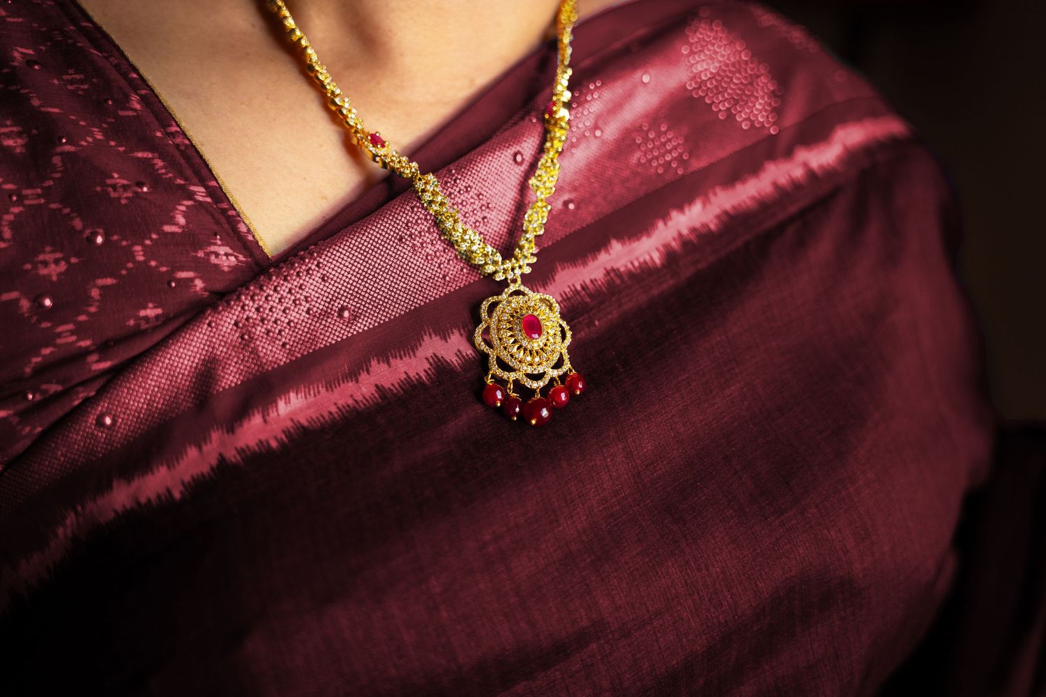 Close-up of a gold and ruby pendant necklace worn over a deep burgundy silk saree.