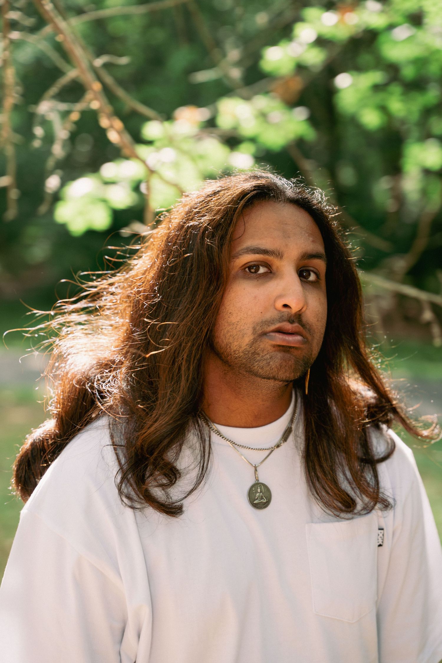 Young man with long brown hair wearing a white shirt and pendant necklace poses outdoors among green trees.