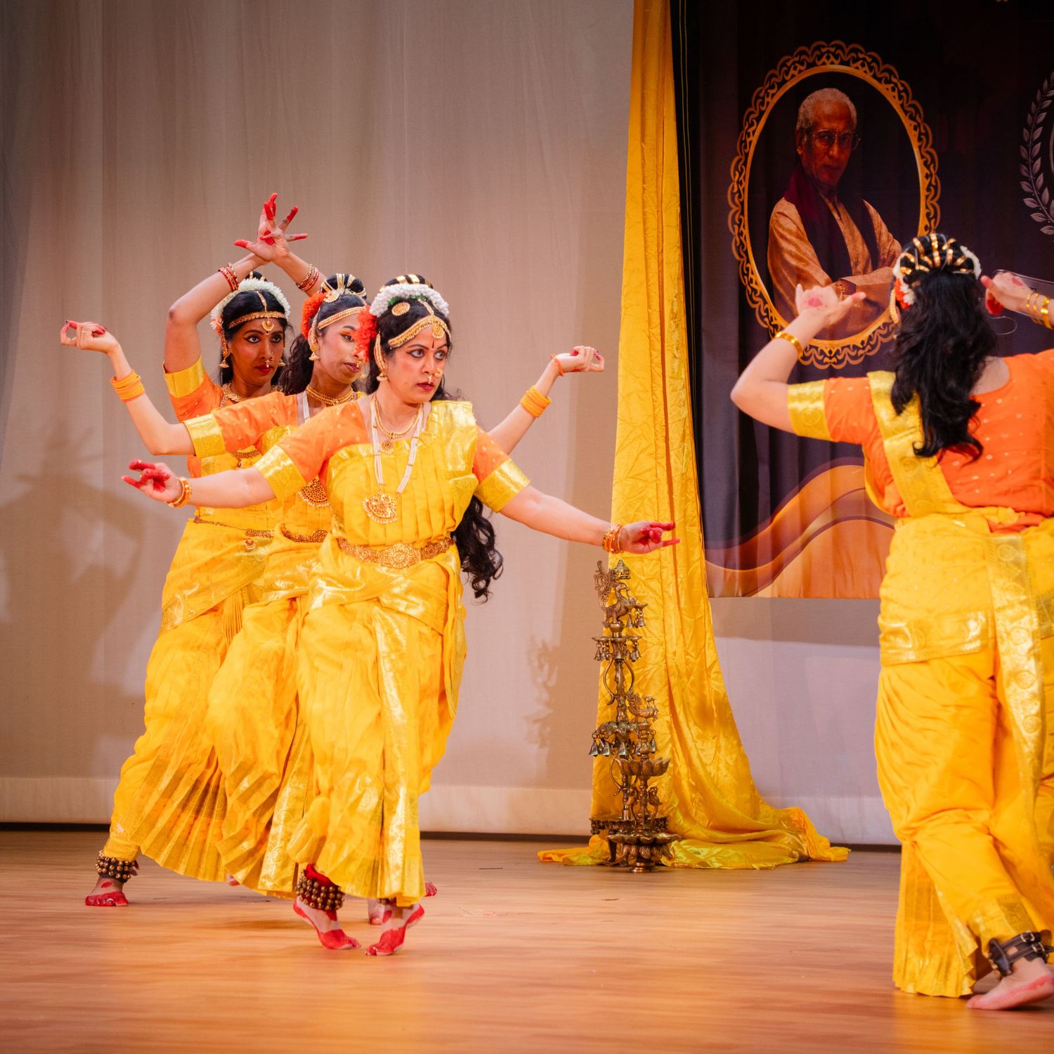 Indian classical dancers in vibrant yellow costumes perform Bharatanatyam on stage during a cultural event.