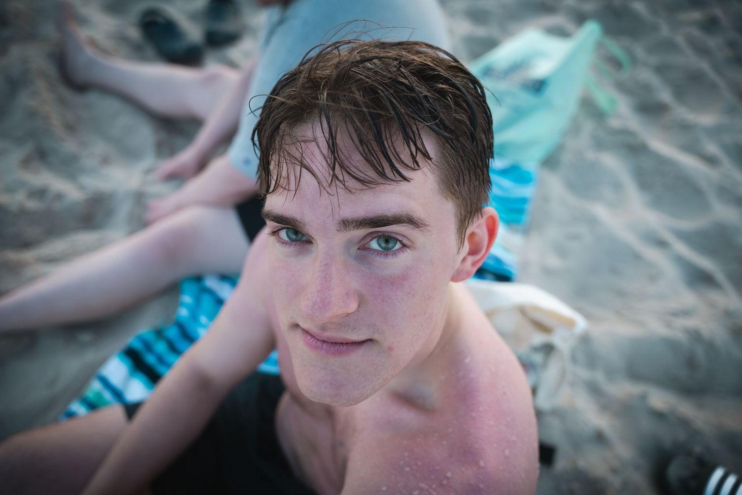 A young man with blue eyes relaxes on a sandy beach looking up at the camera.