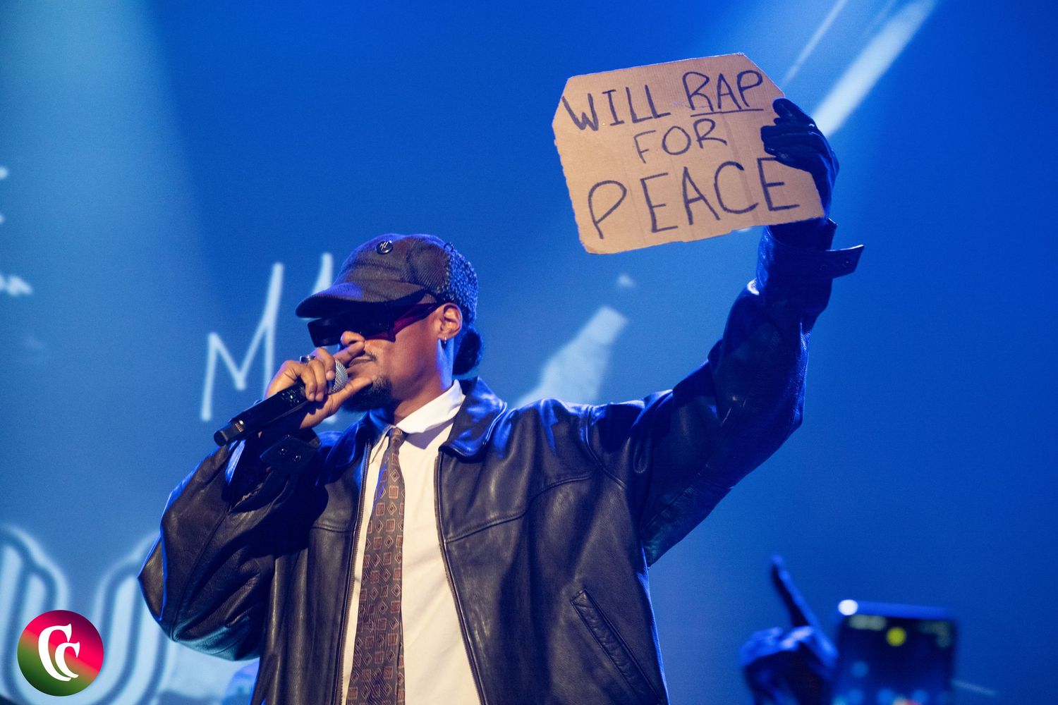 Rapper on stage holding a cardboard sign reading Will Rap For Peace under bright lights.
