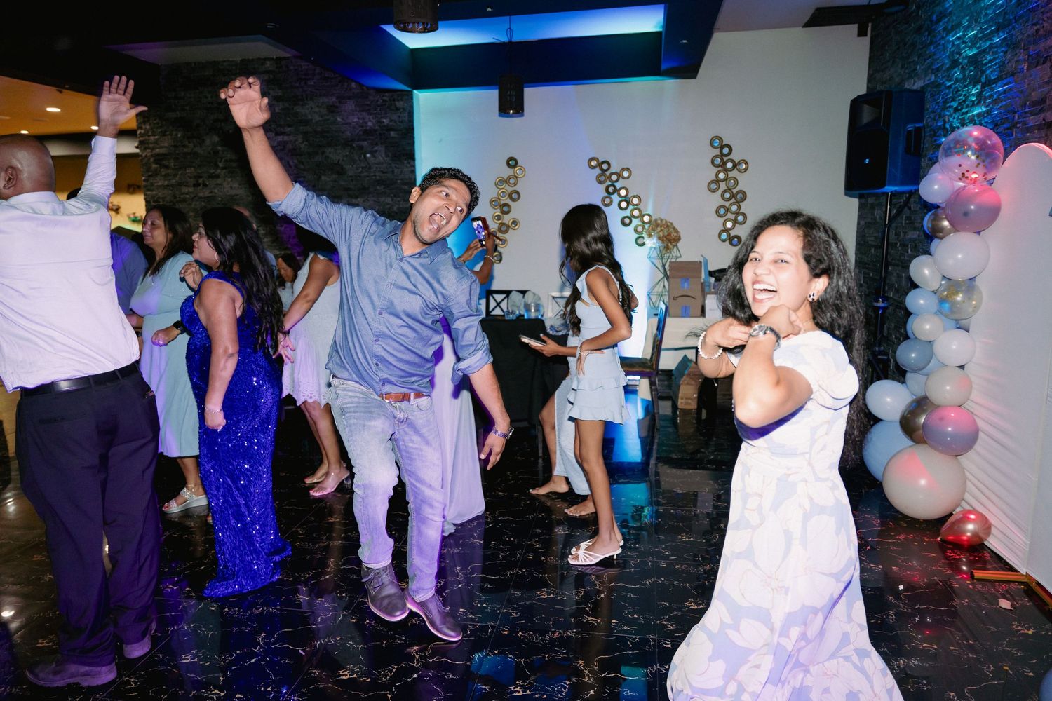 Guests in blue and white outfits dance energetically on a decorated dance floor at a celebration.