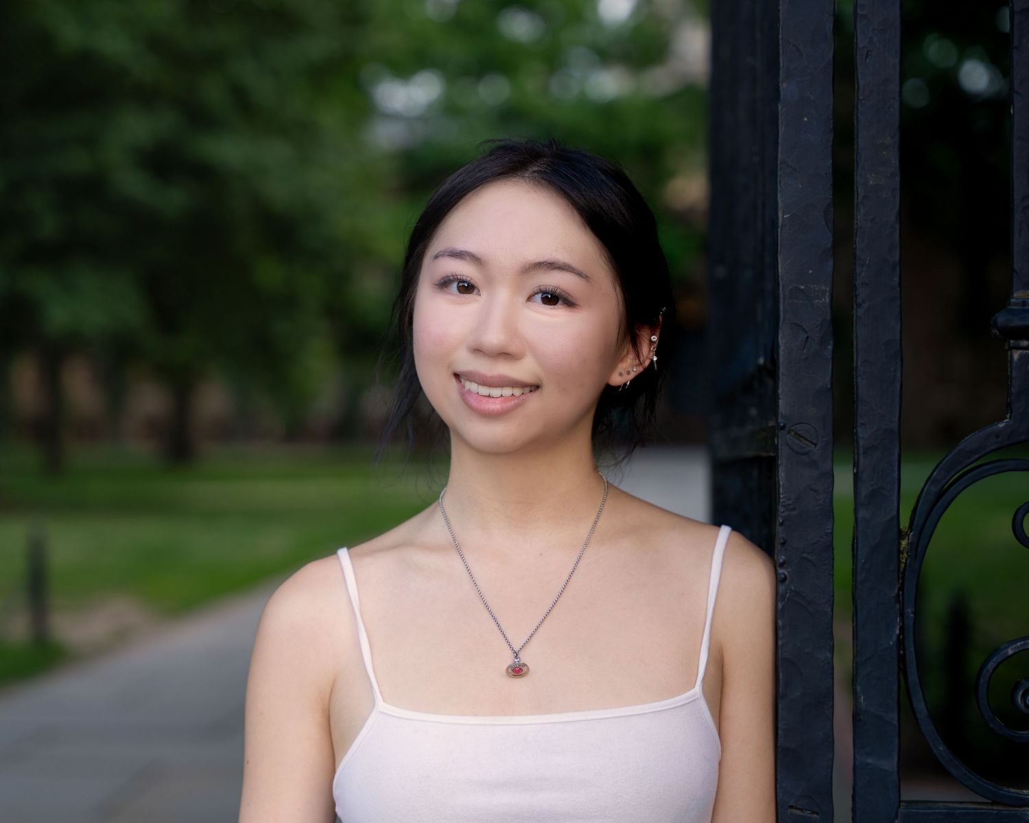 A young woman smiles softly while leaning against a black iron gate in a park.