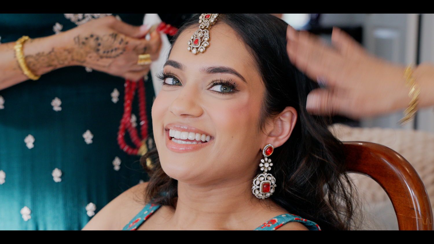 Smiling South Asian bride getting hair styled, wearing ornate red and gold chandelier earrings and traditional jewelry.