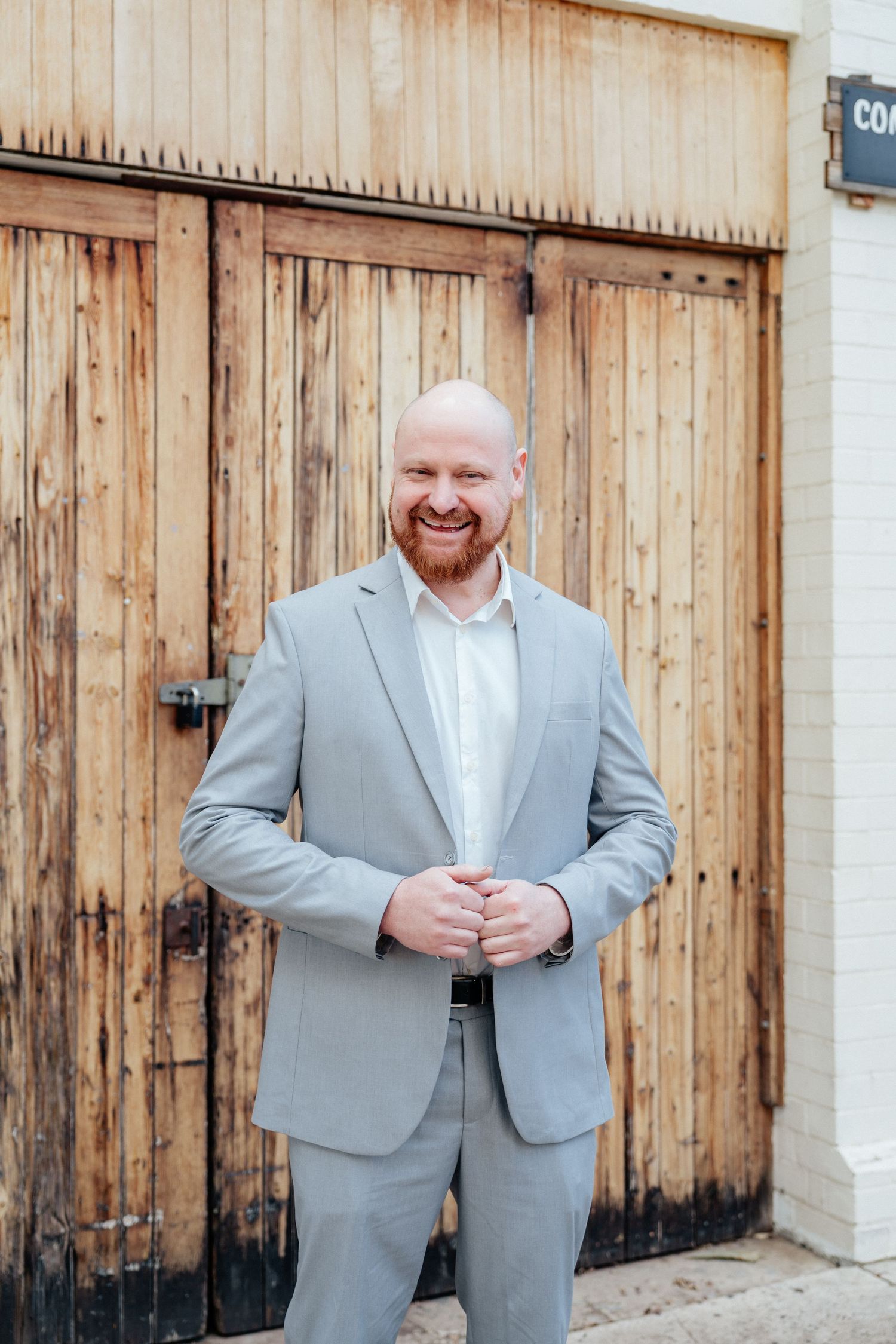 Smiling bald groom in a light grey suit standing in front of rustic wooden doors.