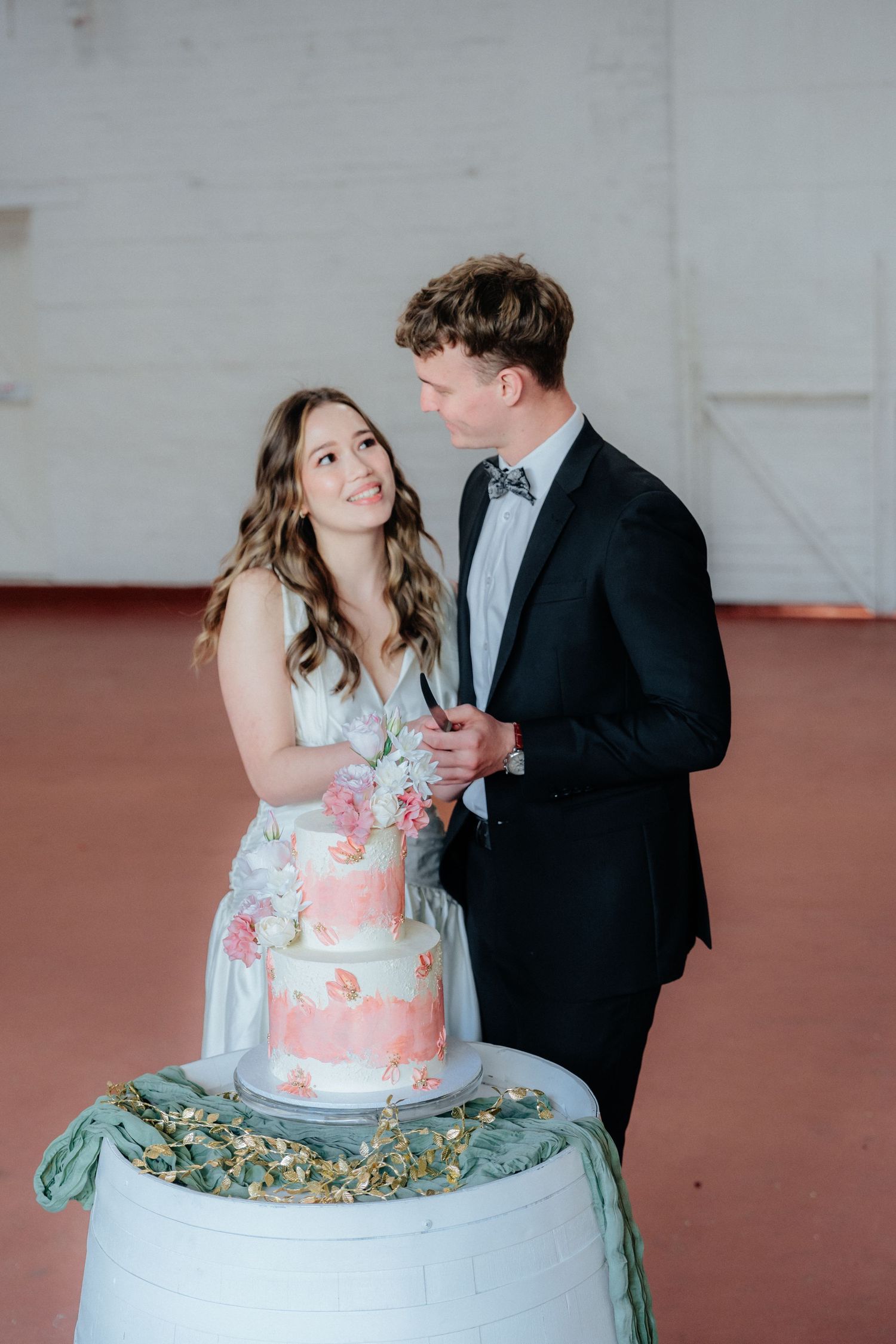 Bride and groom cutting a floral wedding cake together, smiling at each other in an elegant venue.