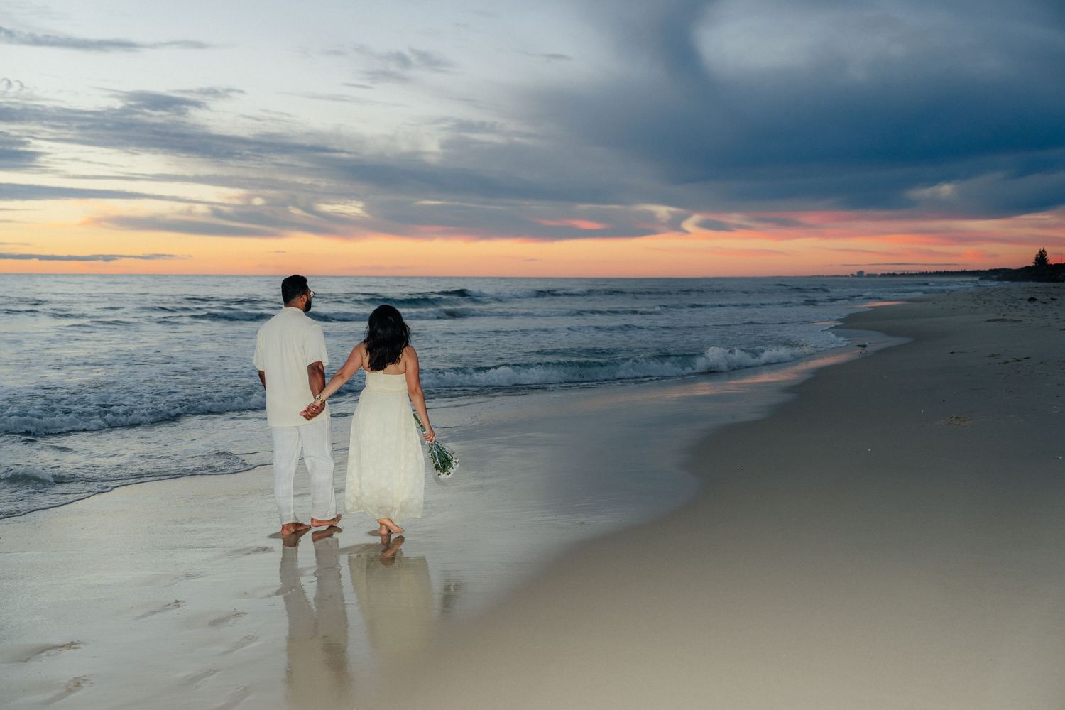 Couple holding hands gazing at dramatic colorful sunset sky over ocean shoreline.
