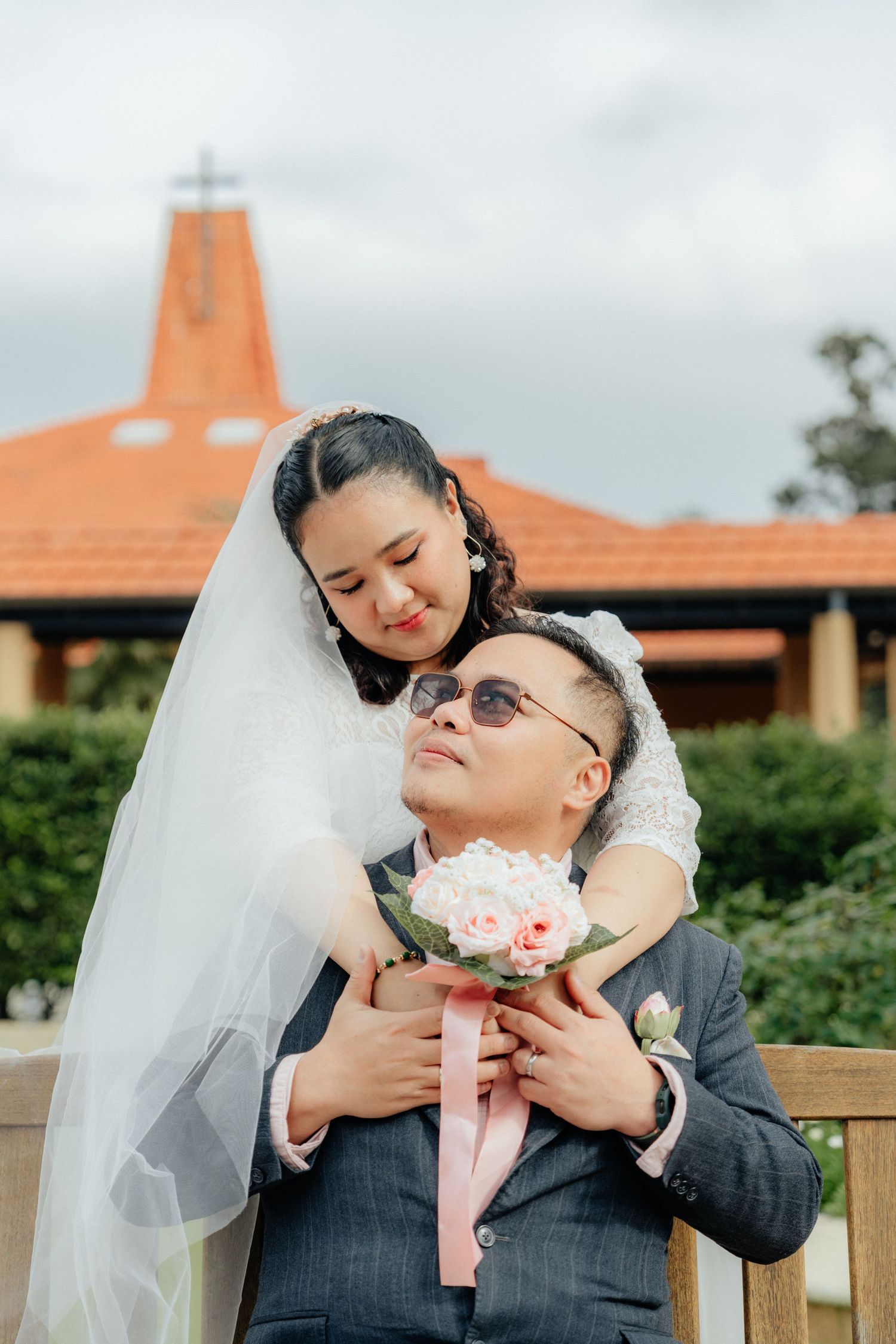 A bride and groom embrace outdoors in front of a church with a cross on the roof.