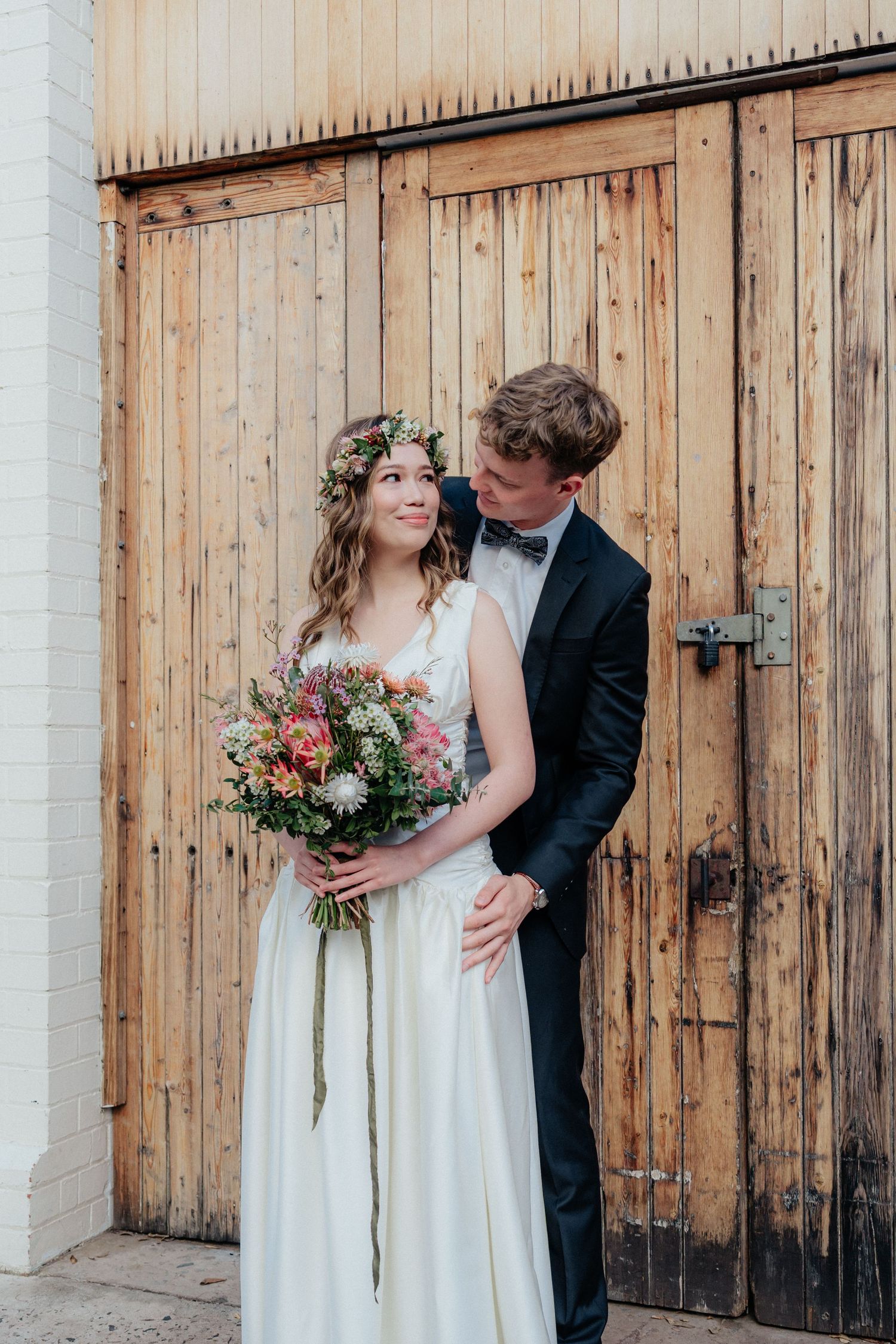 Bride wearing flower crown holding bouquet embraced by groom in black tuxedo before rustic wooden doors.