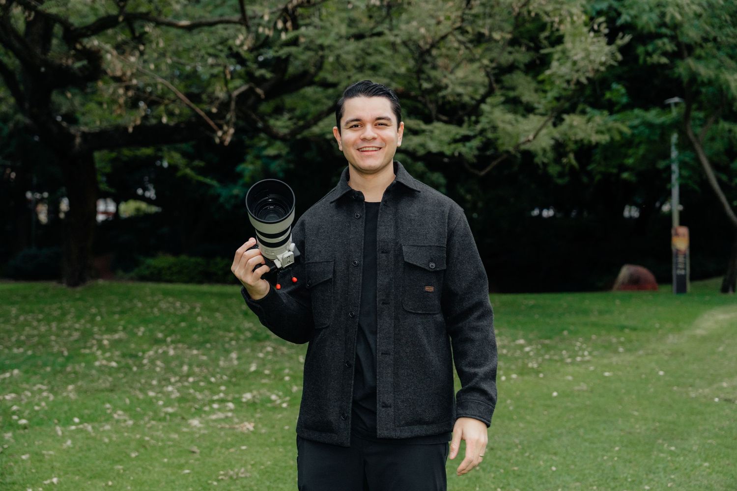 Professional wedding photographer in black outfit holding camera in a green park setting.