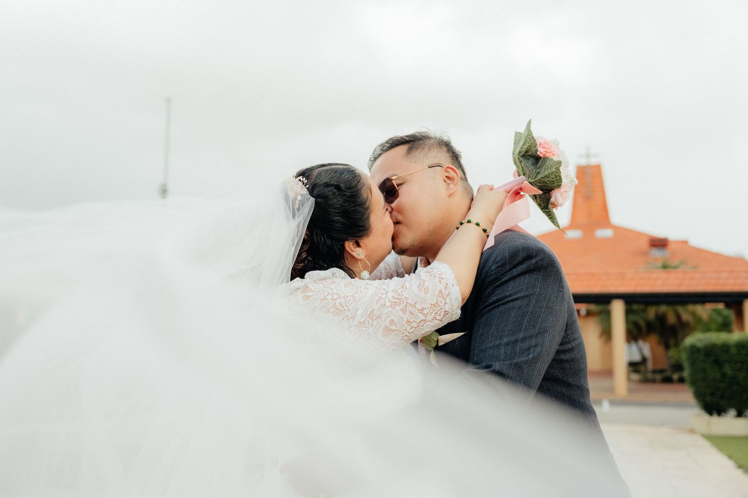 Bride and groom sharing a kiss with wedding veil flowing in the wind outdoors.