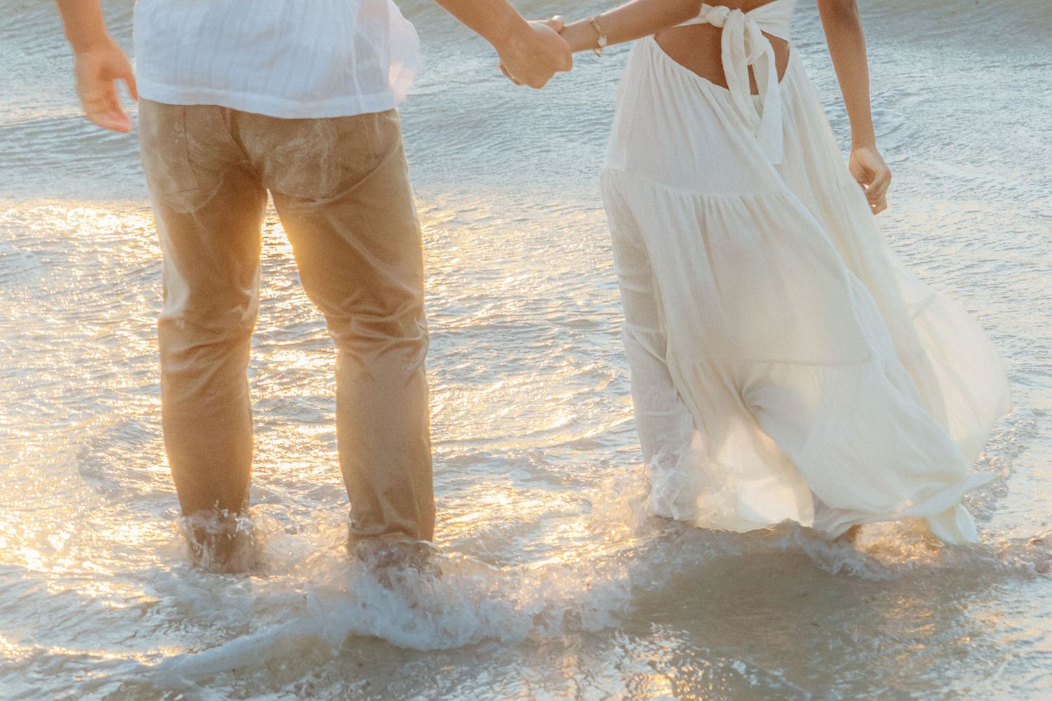 Couple holding hands walking through ocean waves at sunset, wearing white dress and tan pants.