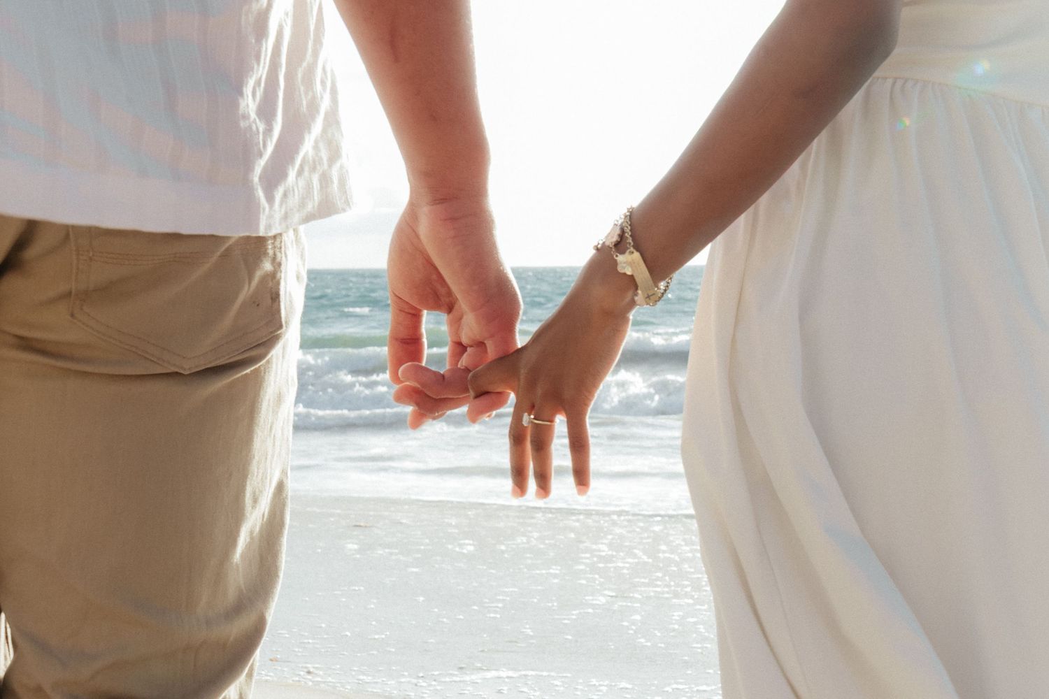Engaged couple holding hands on beach, woman displaying diamond ring, ocean behind them.