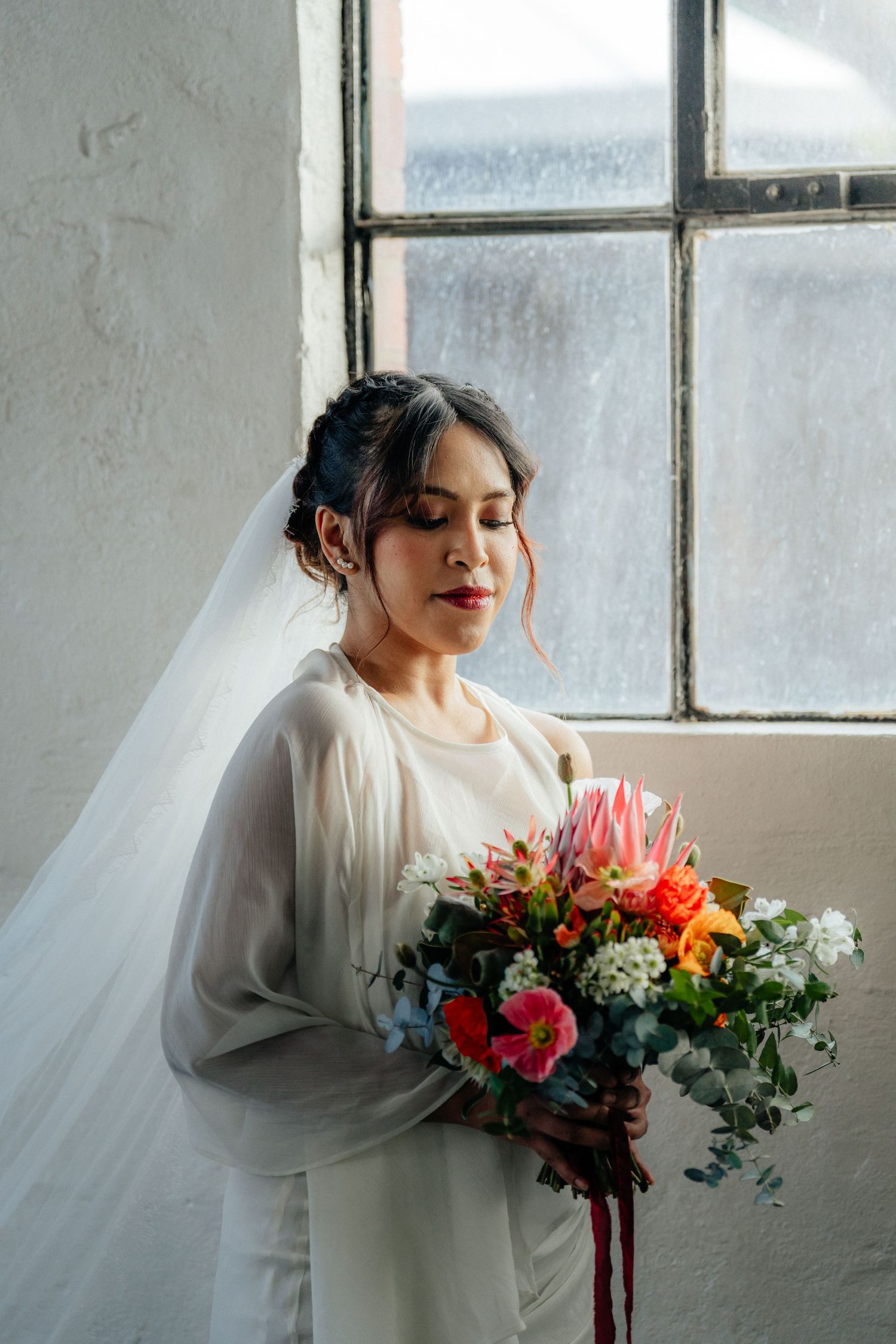 Bride in white gown holding a colorful bouquet with protea flowers by a window.