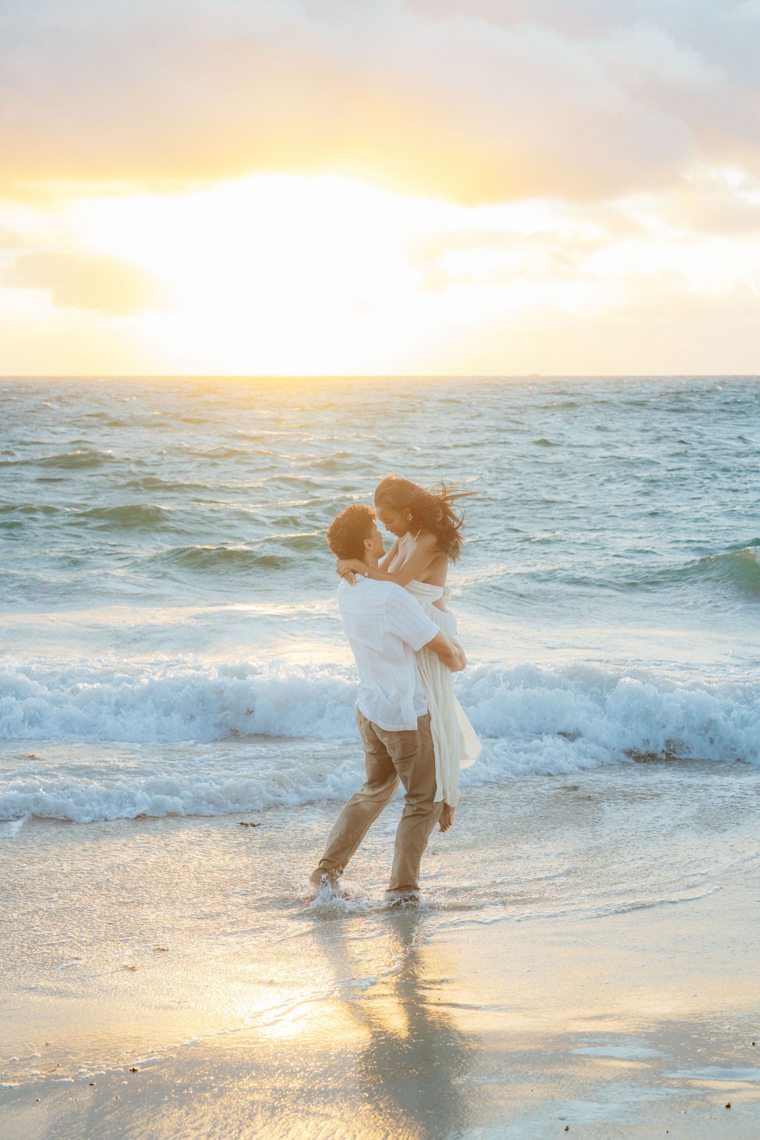 Couple embracing at sunset on a beach with golden waves crashing behind them.