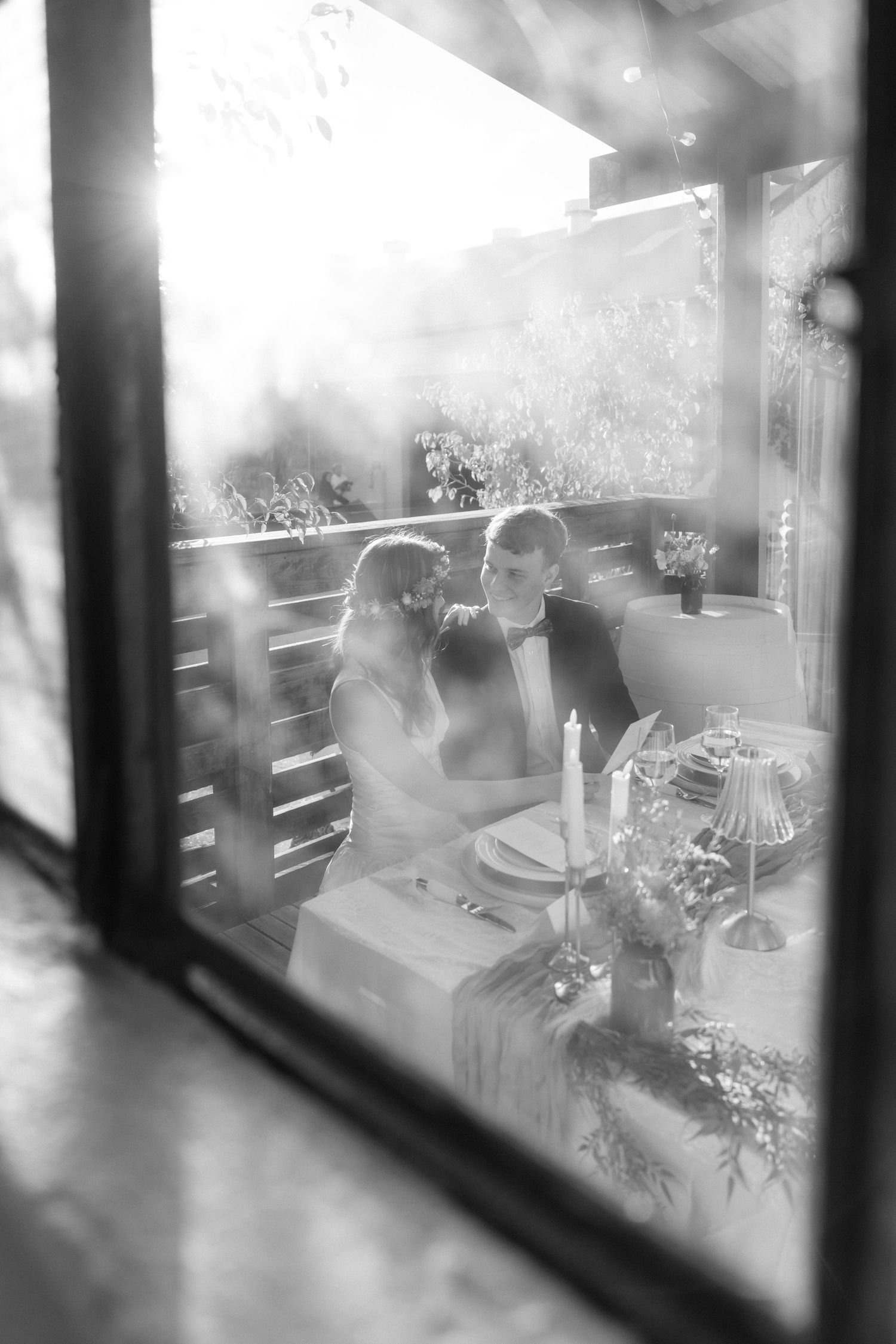 Black and white romantic photo of newlyweds sitting at decorated wedding table, viewed through a window.