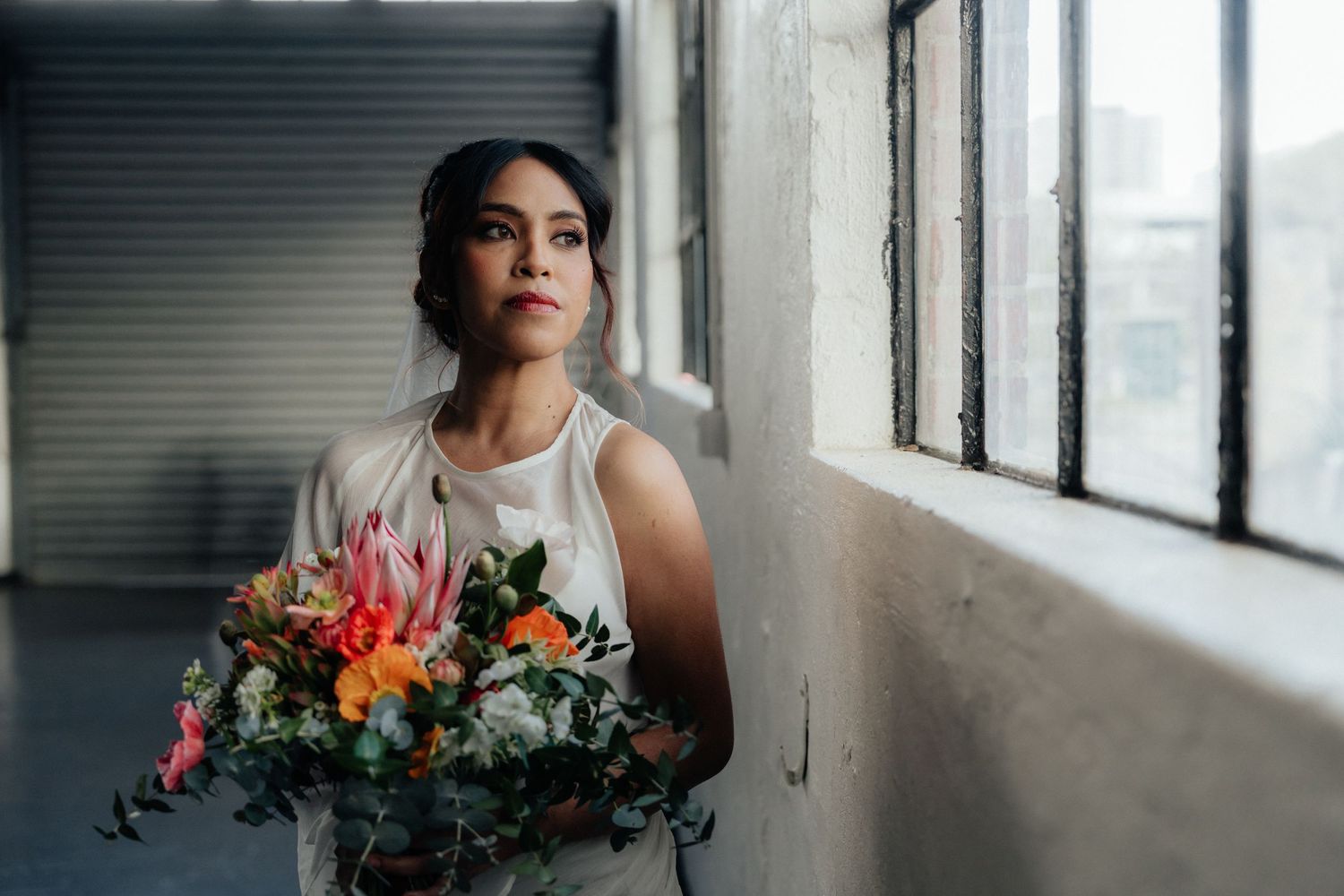 Elegant bride holding a vibrant tropical bouquet standing by industrial windows in soft light.
