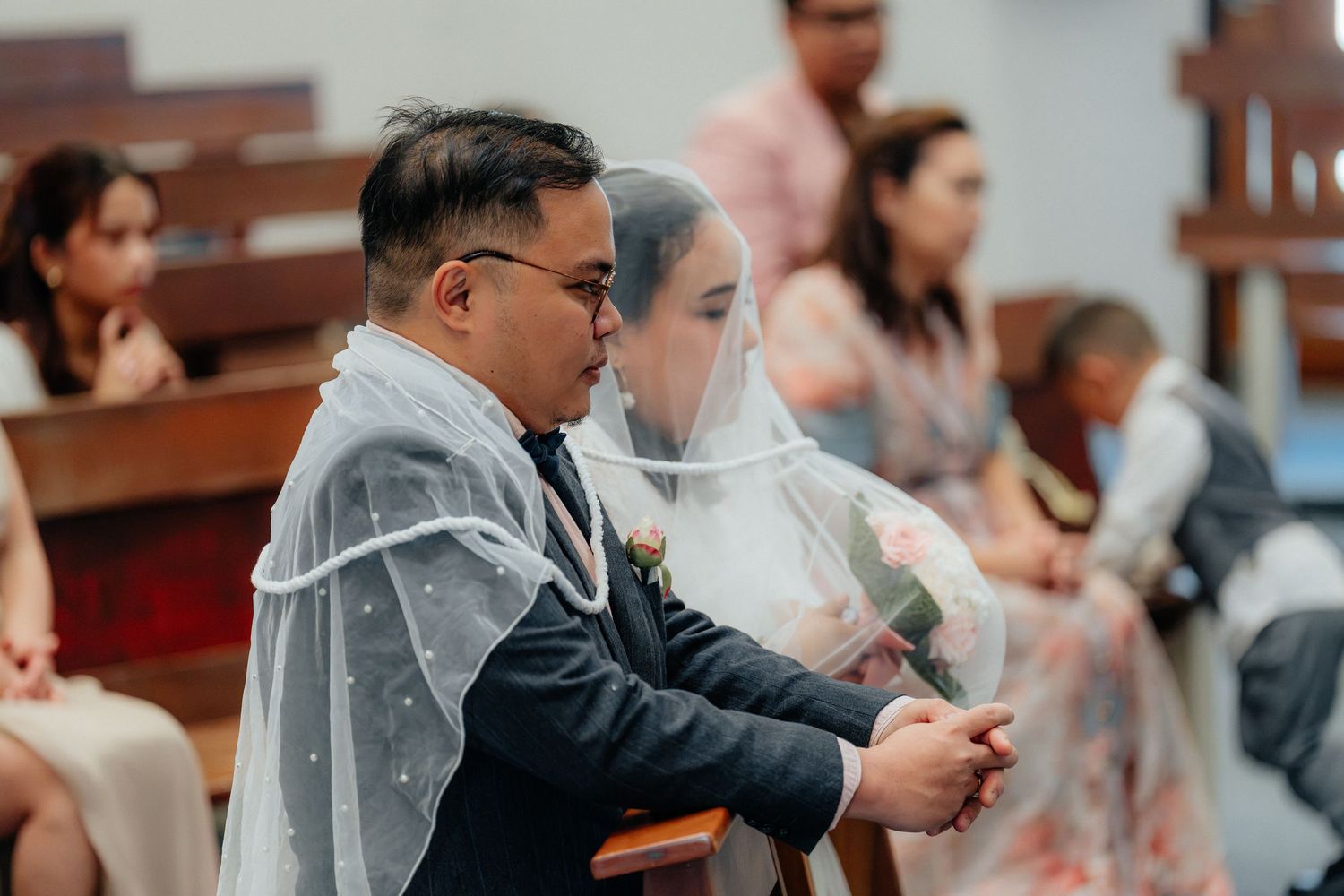 A bride and groom sit together under a wedding veil during a church ceremony.