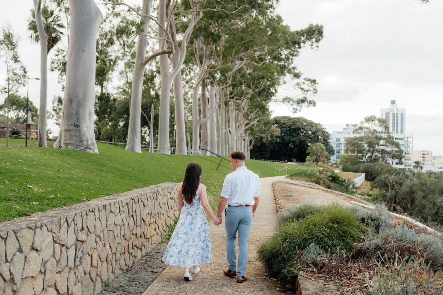 Engaged couple walking hand in hand along a stone path in a scenic park.