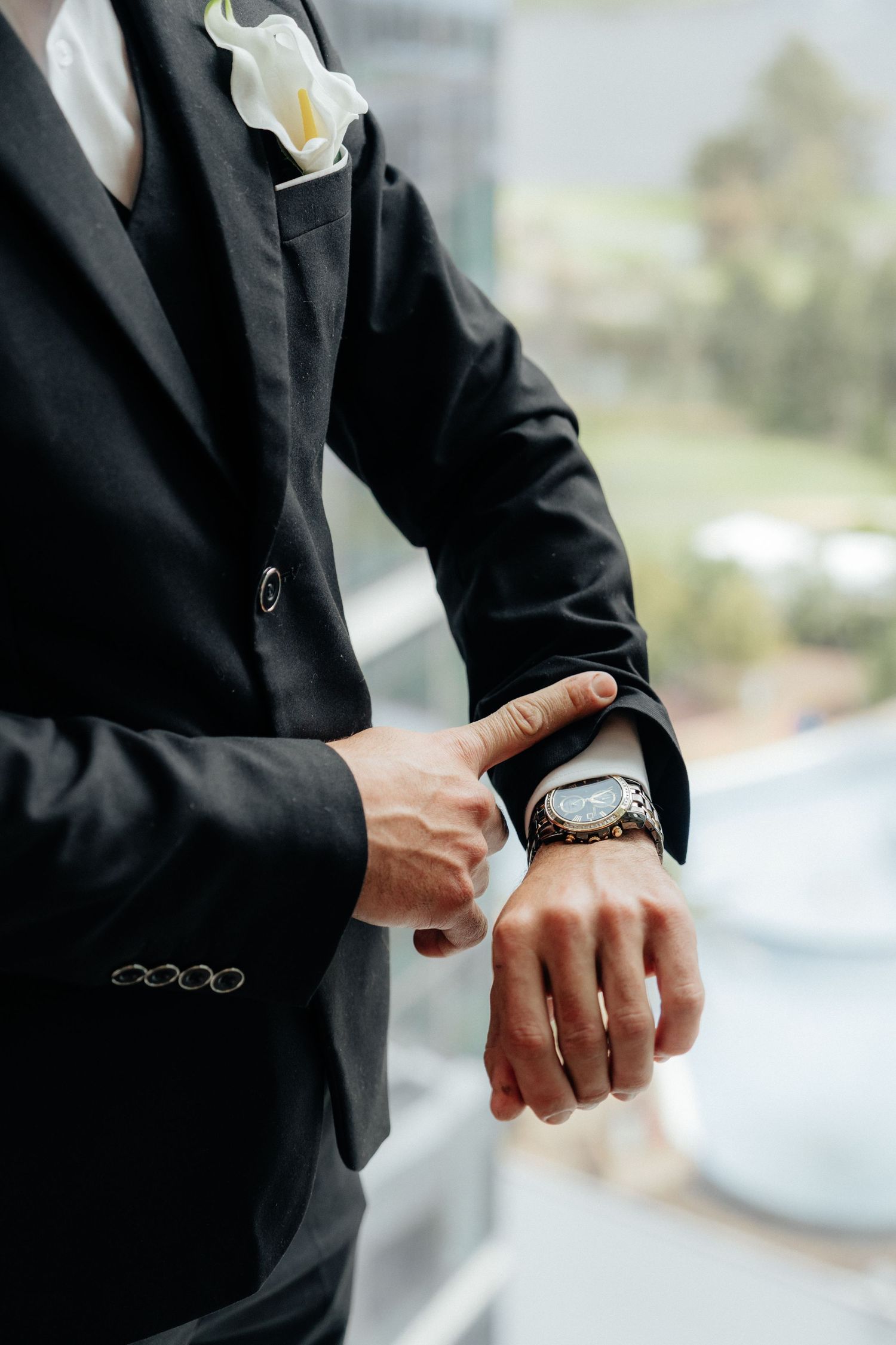Groom in black suit adjusting wristwatch with white calla lily boutonniere, wedding day preparation.
