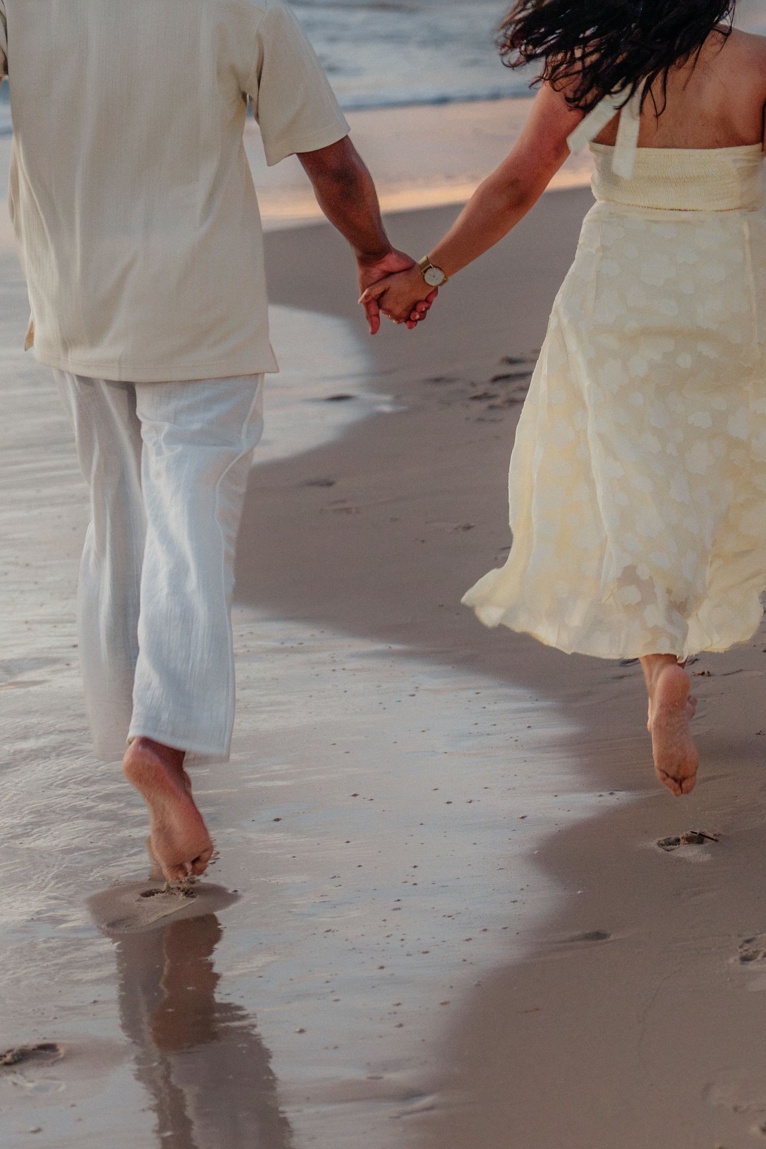 Barefoot couple holding hands walking along wet shoreline, captured from behind at beach.