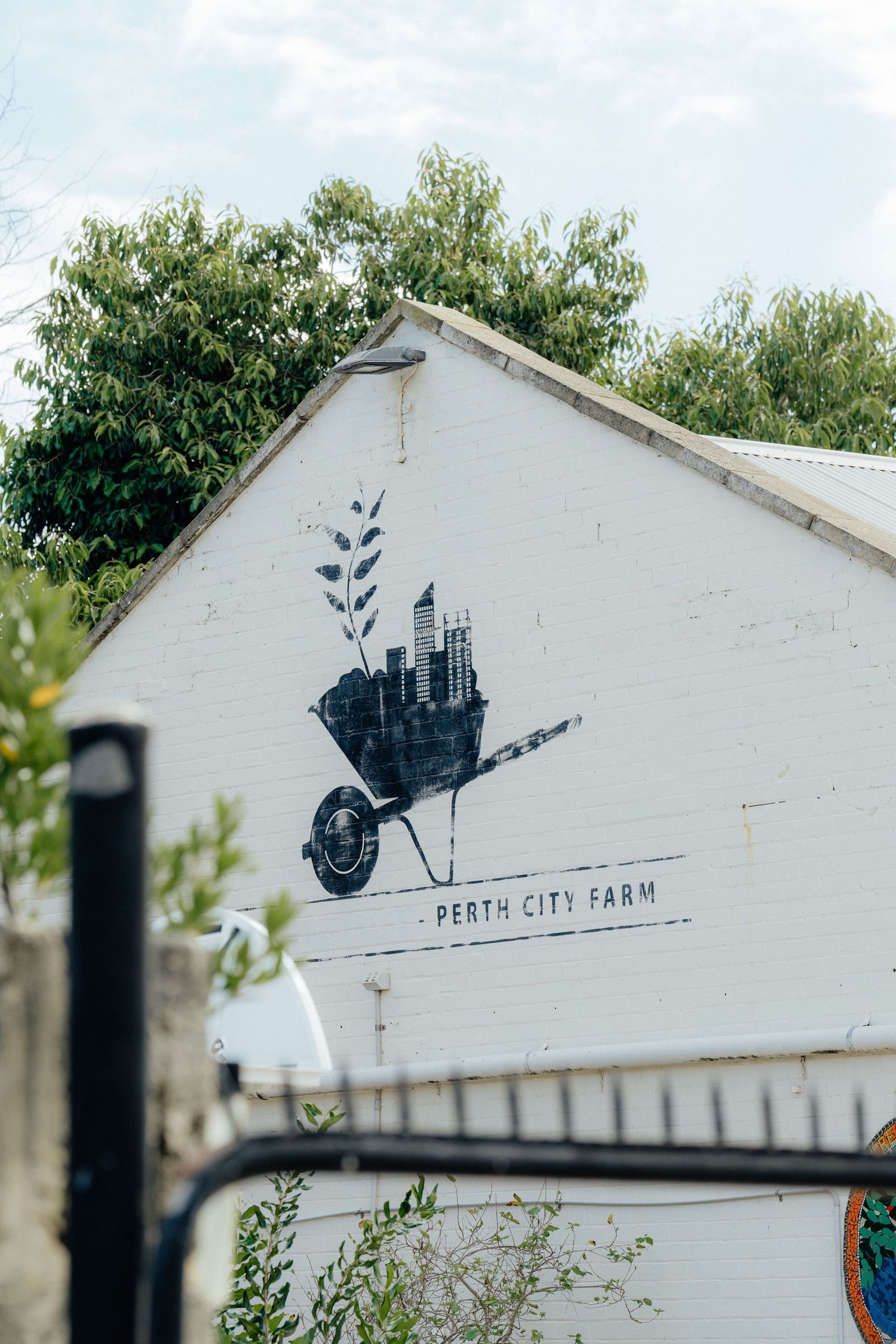 Perth City Farm logo mural painted on white building exterior wall with wheelbarrow cityscape art.