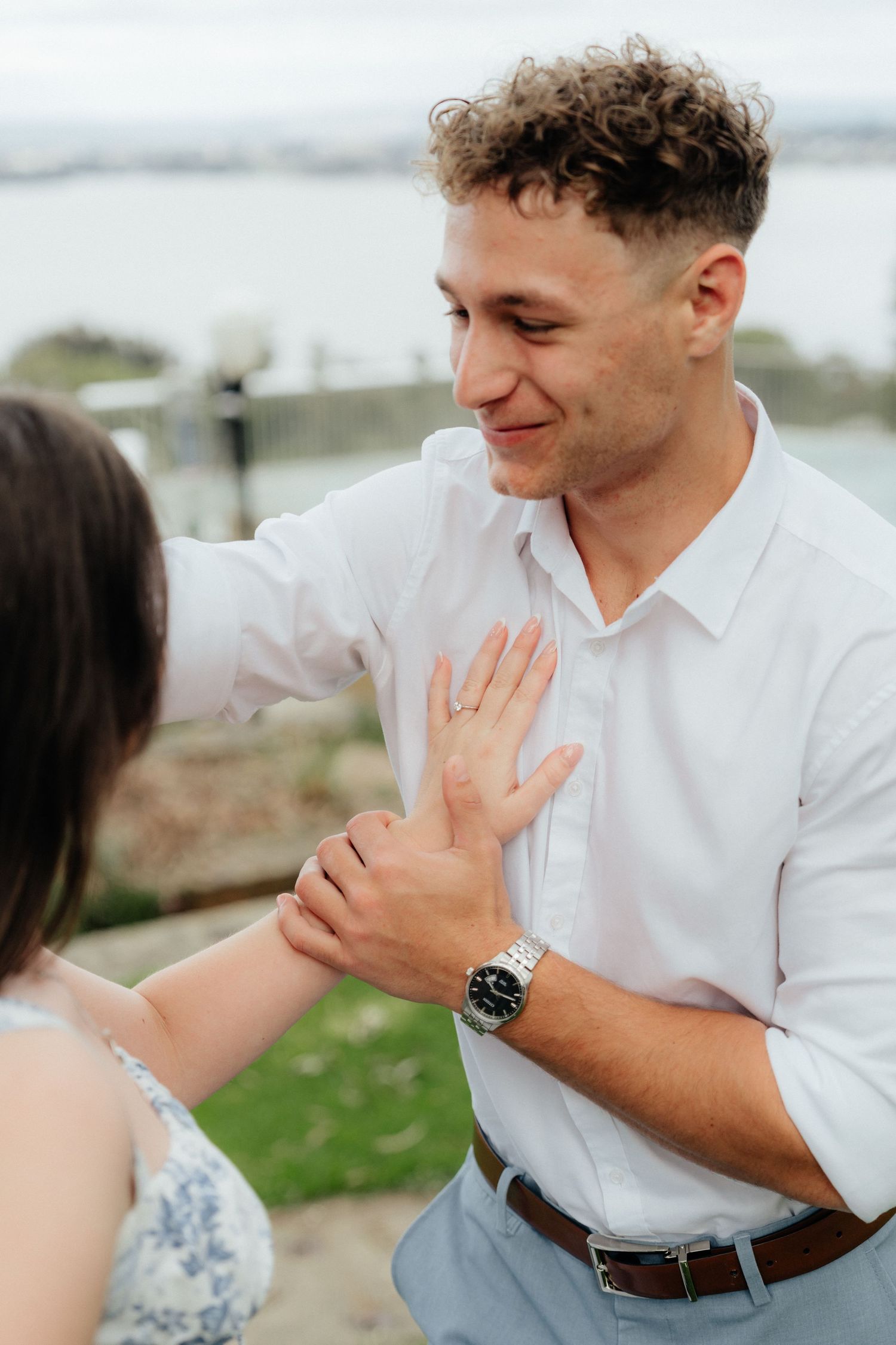 Smiling groom in white shirt holds his partners hand showing engagement ring with city backdrop.