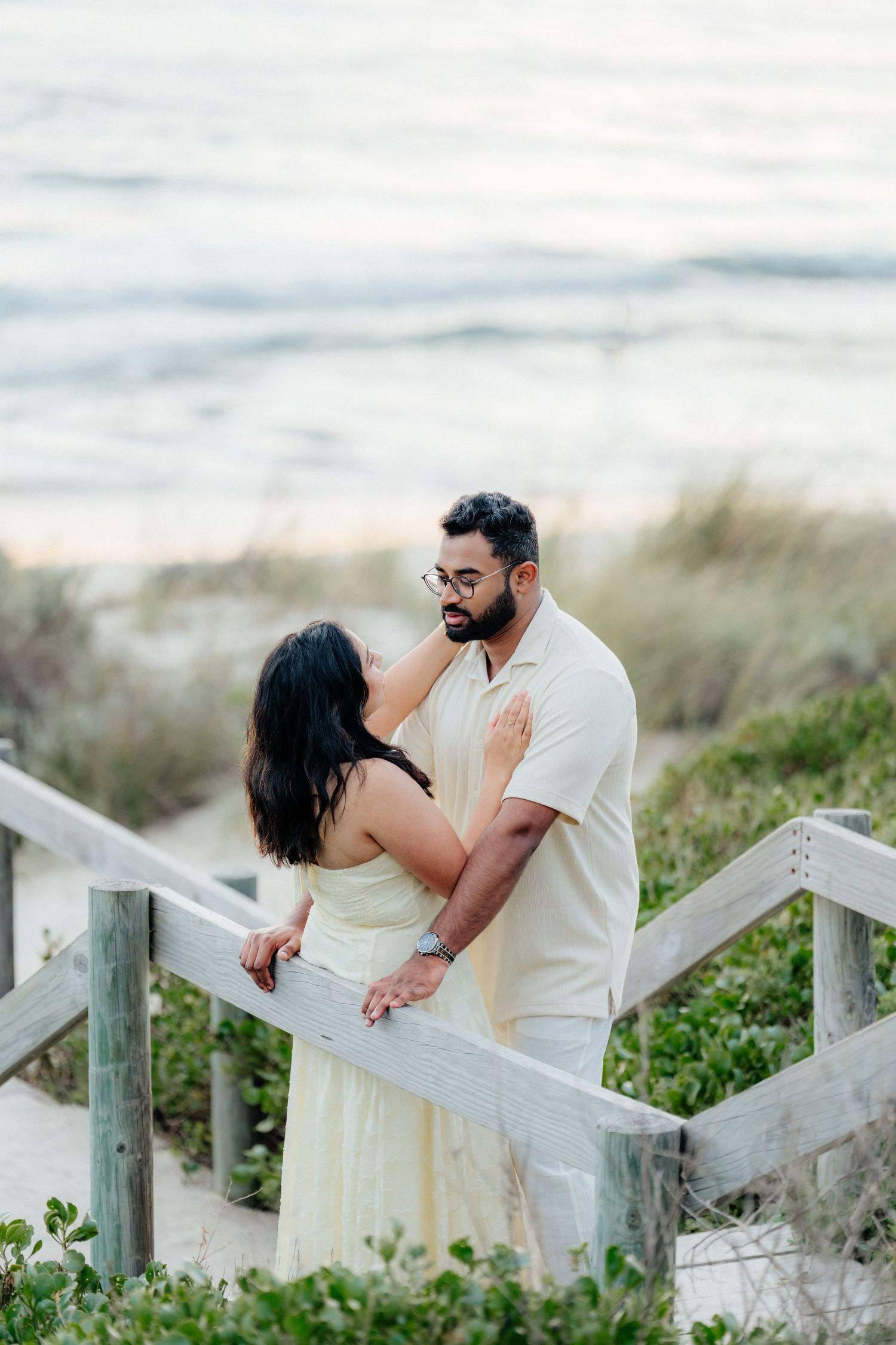 Couple sharing a romantic moment on wooden beach boardwalk stairs with ocean waves in the background.