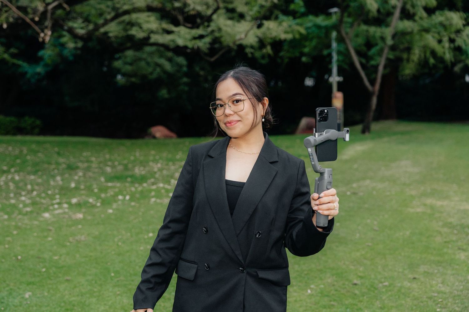 Young Asian female videographer in black blazer holding smartphone gimbal stabilizer in a park.