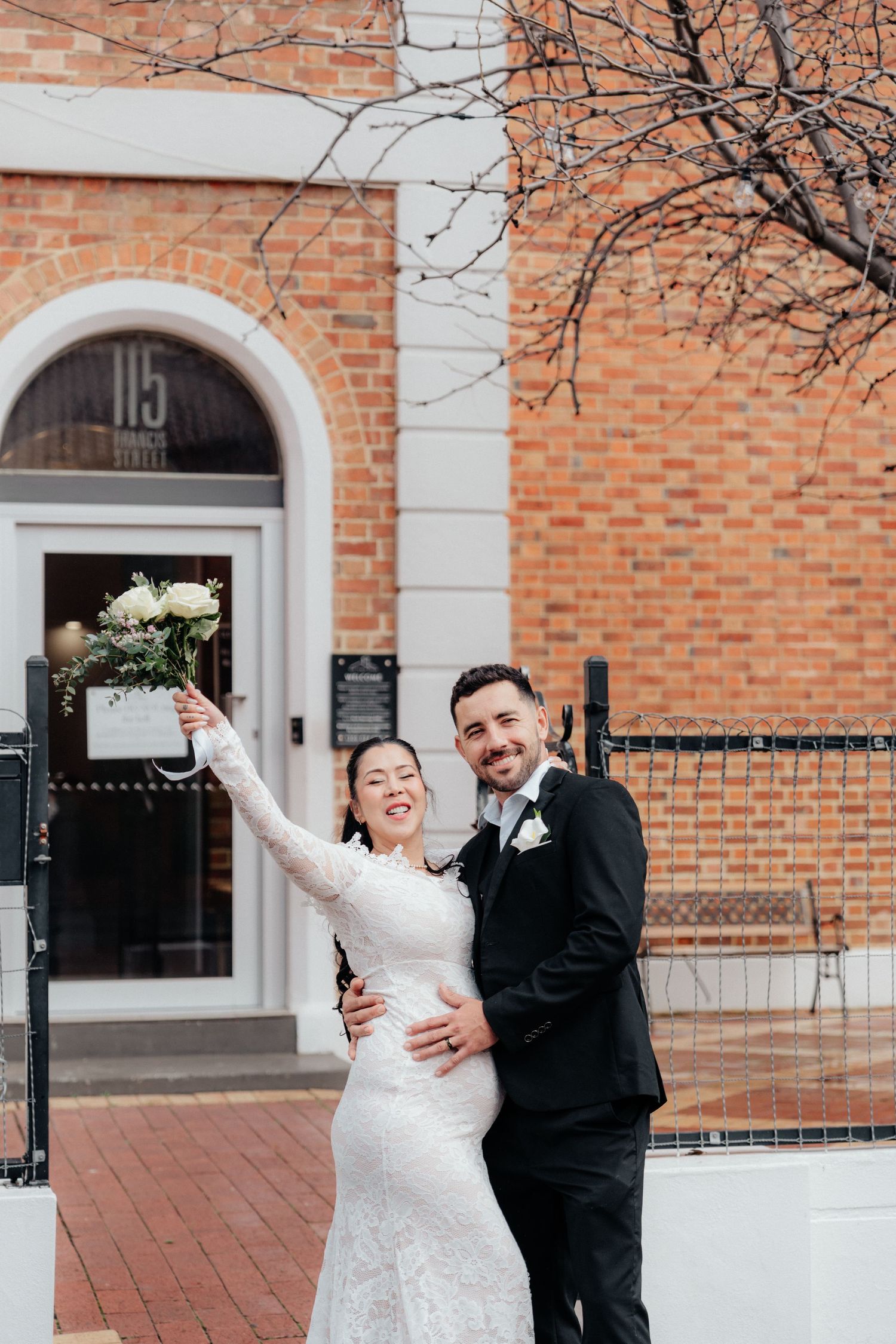 Happy bride and groom pose together outside brick building, bride raising bouquet in celebration after wedding ceremony.