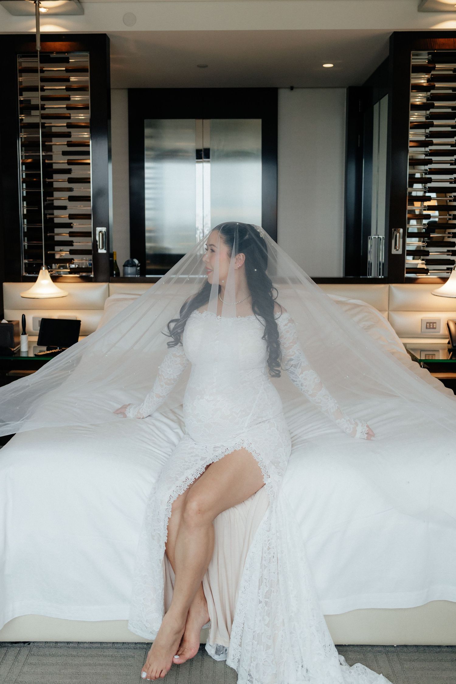Elegant bride in white lace gown and veil sits gracefully on white hotel bed during wedding preparations.
