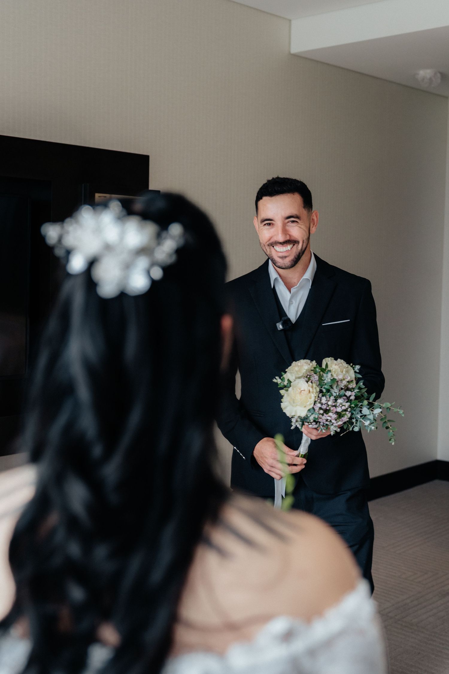 Smiling groom in black suit presents white flower bouquet to bride during wedding first look moment indoors.