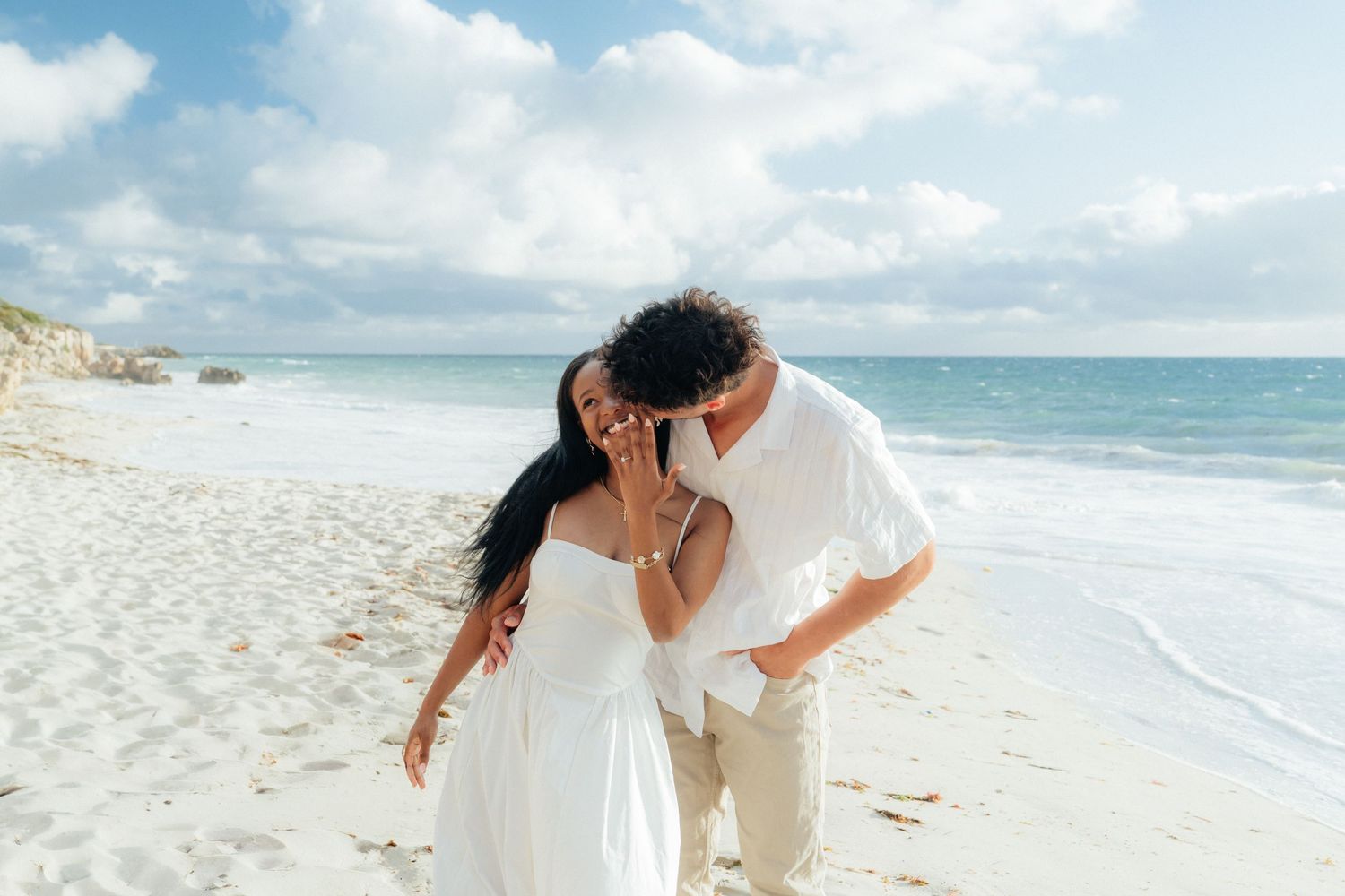 Man kissing womans cheek as she smiles on a bright white sandy beach.