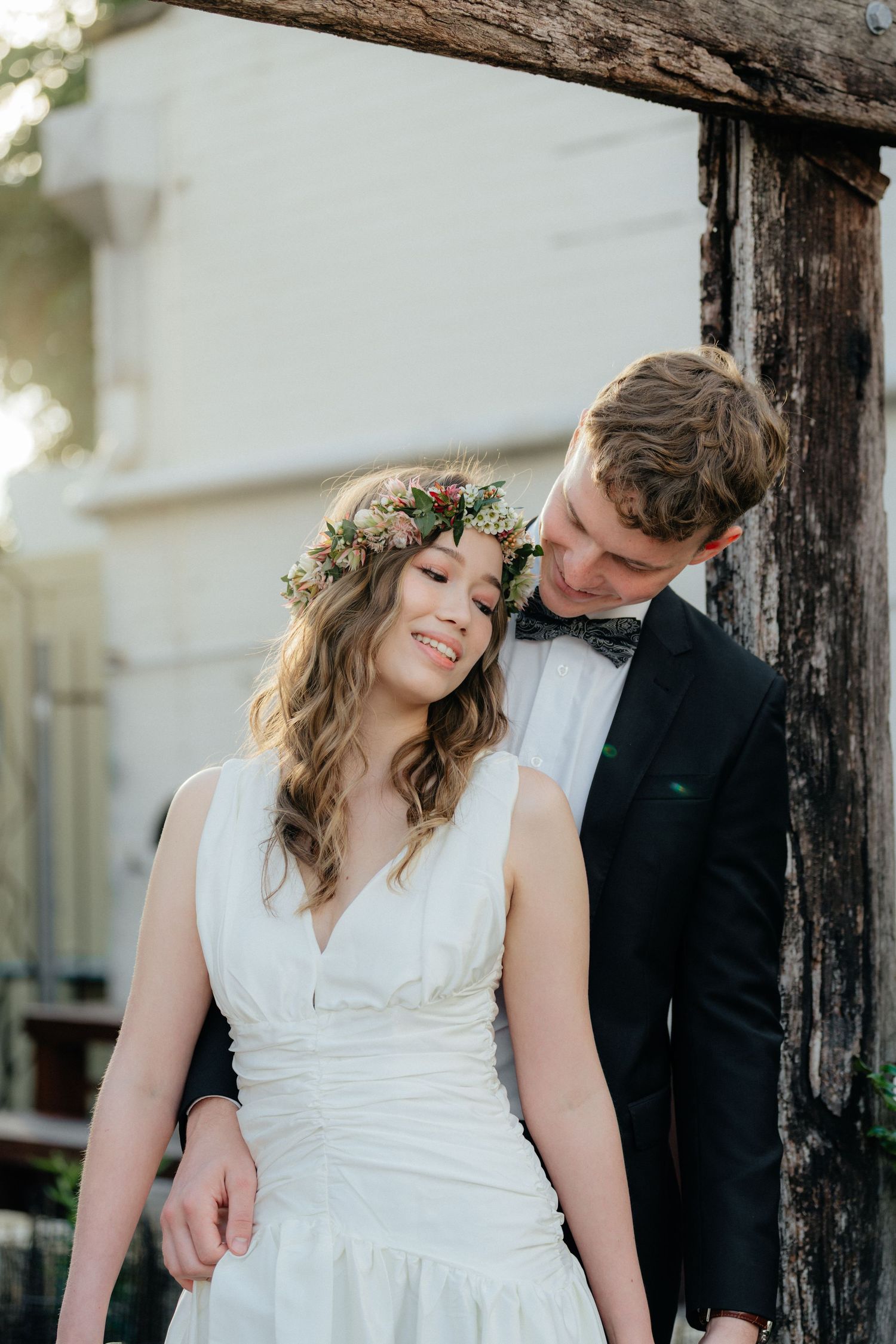 Smiling bride in white dress and flower crown leans against groom in black suit near rustic wood post.