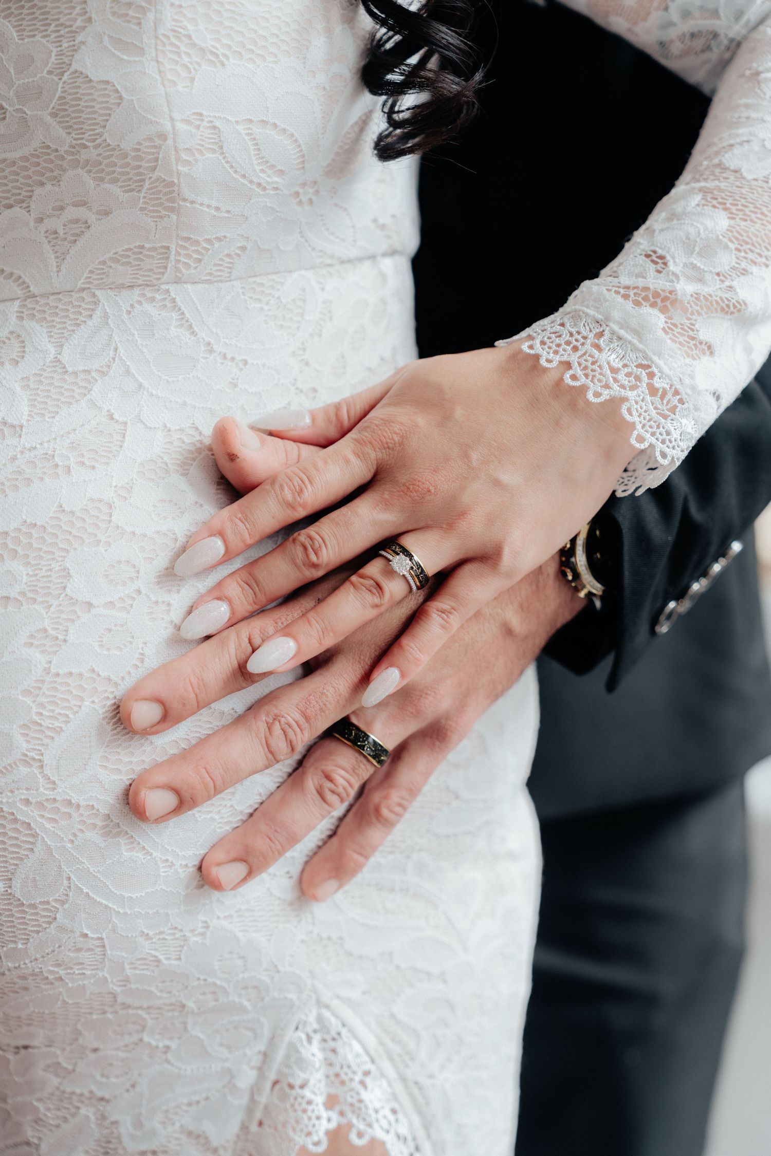 Close-up of bride and groom's hands displaying matching wedding bands over white lace bridal gown.