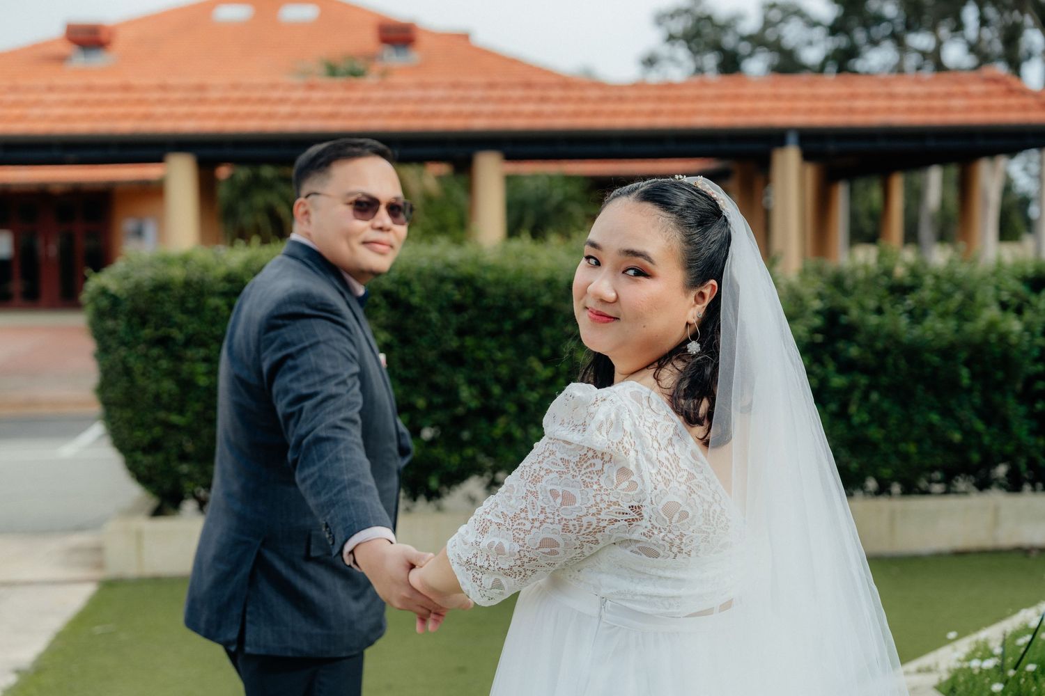 A smiling bride in a white lace gown holds hands with her groom outside a church.