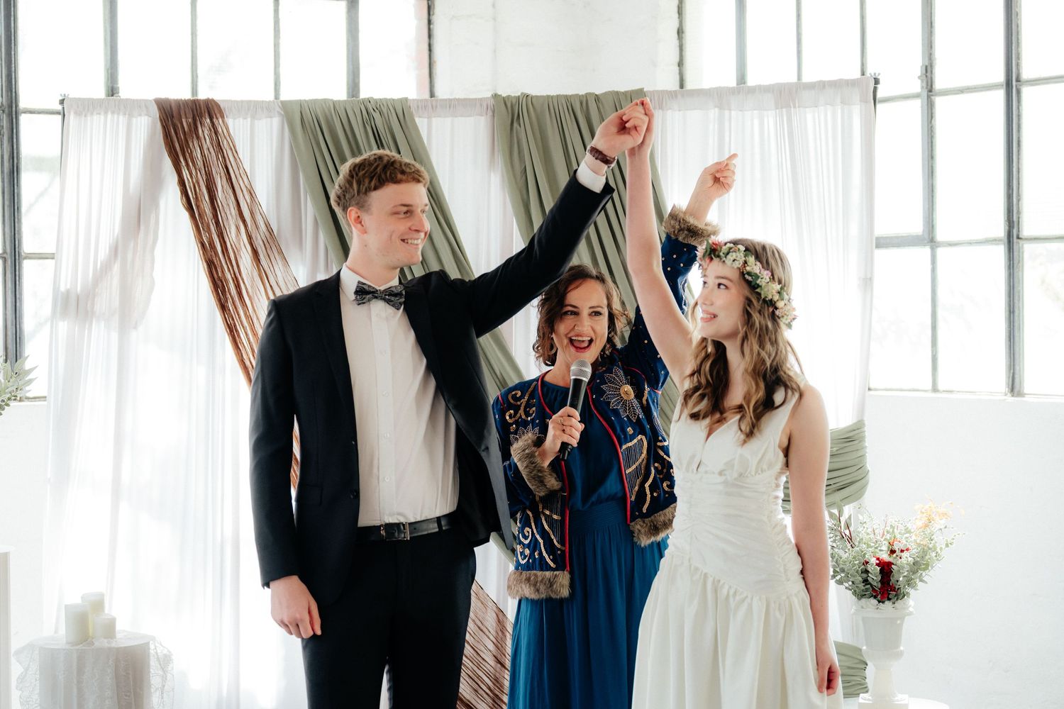 A bride and groom celebrate with raised hands as an officiant joins them at an indoor ceremony.