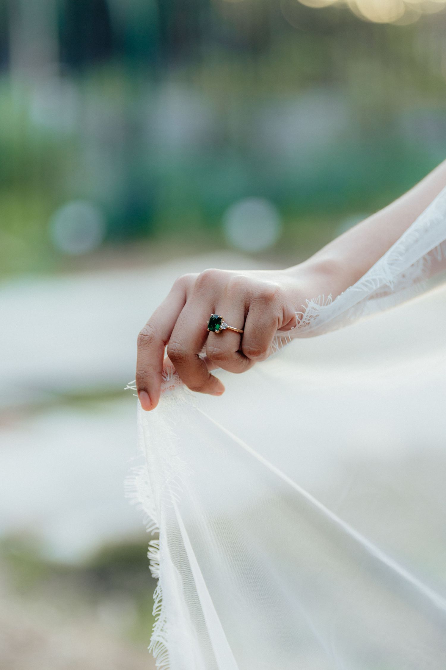 Close-up of brides hand holding white veil with a green emerald engagement ring.