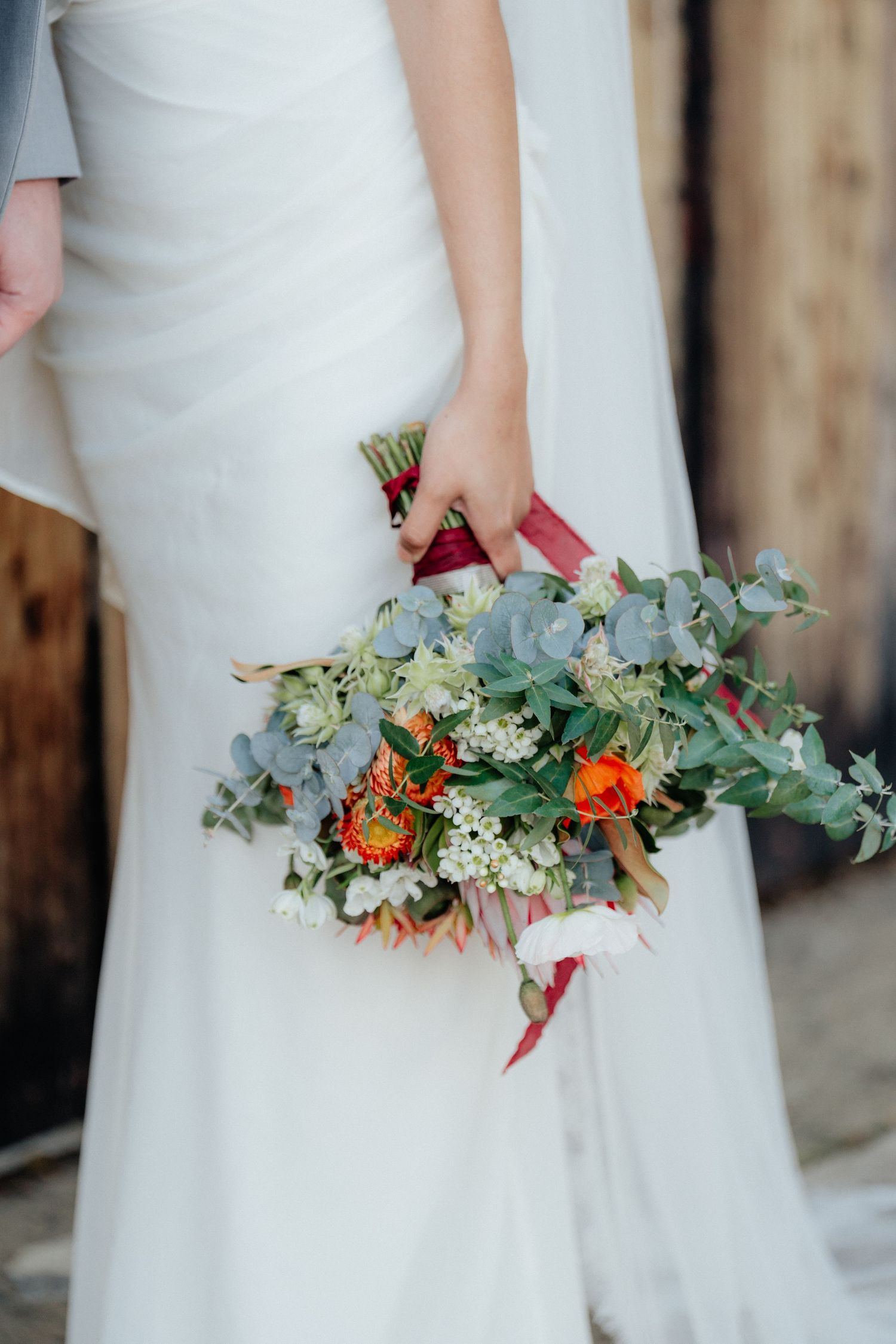 Bride holding a lush wedding bouquet with orange flowers, eucalyptus, and red ribbons.