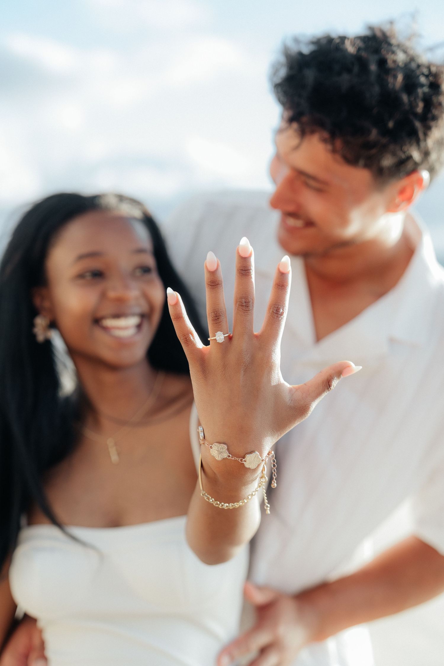 Newly engaged couple showing off diamond engagement ring on the beach in white outfits.