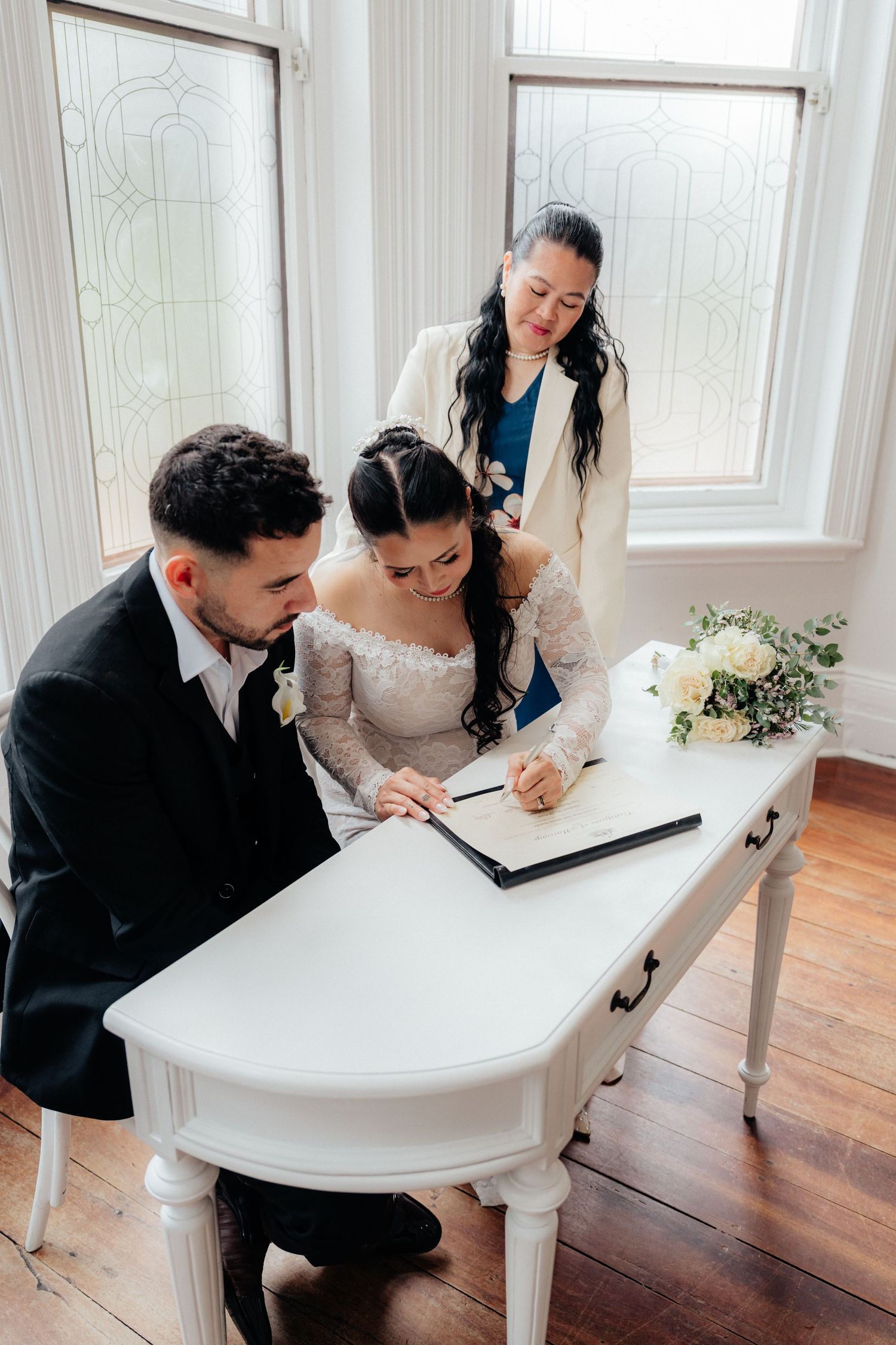 Bride and groom sign wedding register at white desk with officiant witness standing nearby.