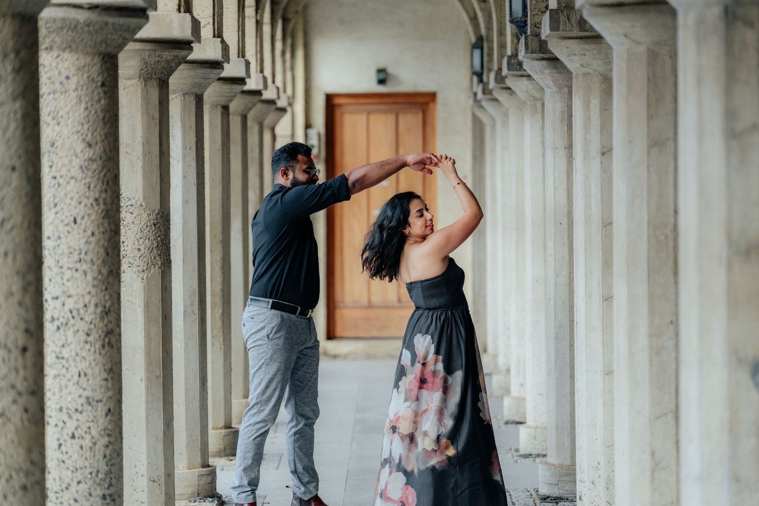 Couple dancing under stone columns of a historic building, woman in a floral gown.