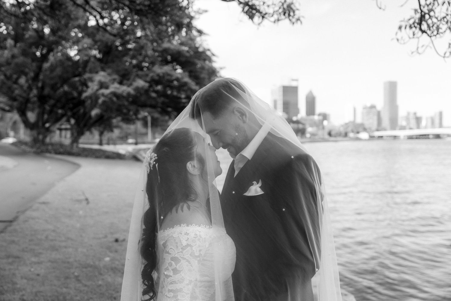 Black and white romantic photo of bride and groom under veil by a city waterfront.