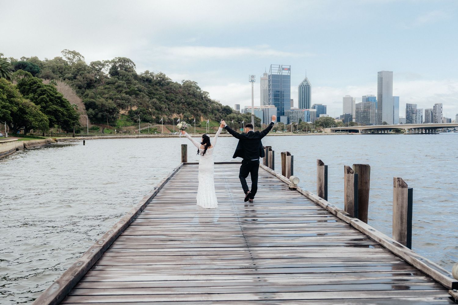 A bride and groom celebrate on a wooden pier with a city skyline in the background.