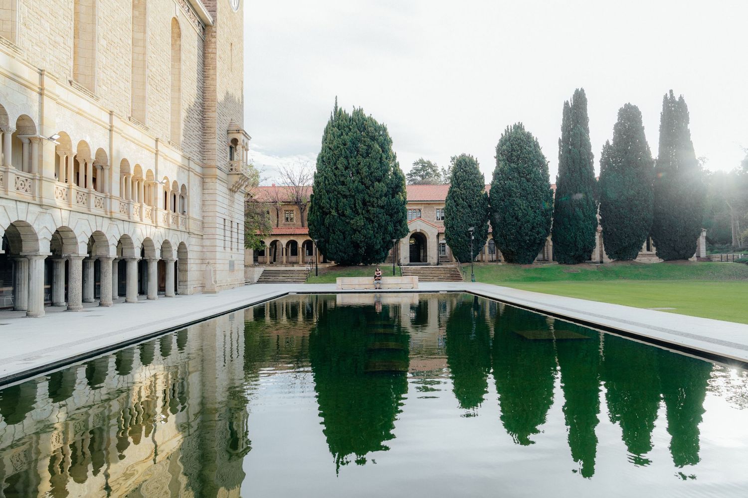 Reflecting pool mirroring trees and historic stone arched building on a cloudy day.