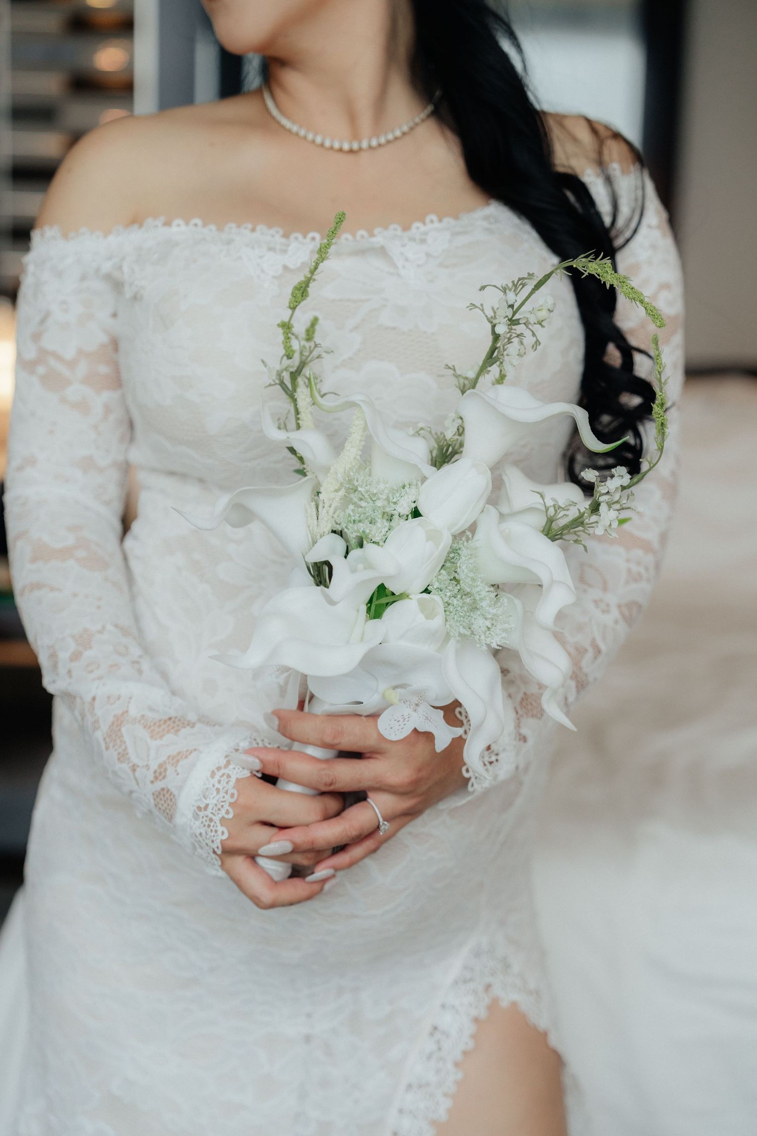 Bride in off-shoulder lace wedding gown holding elegant white calla lily bouquet with pearl necklace.
