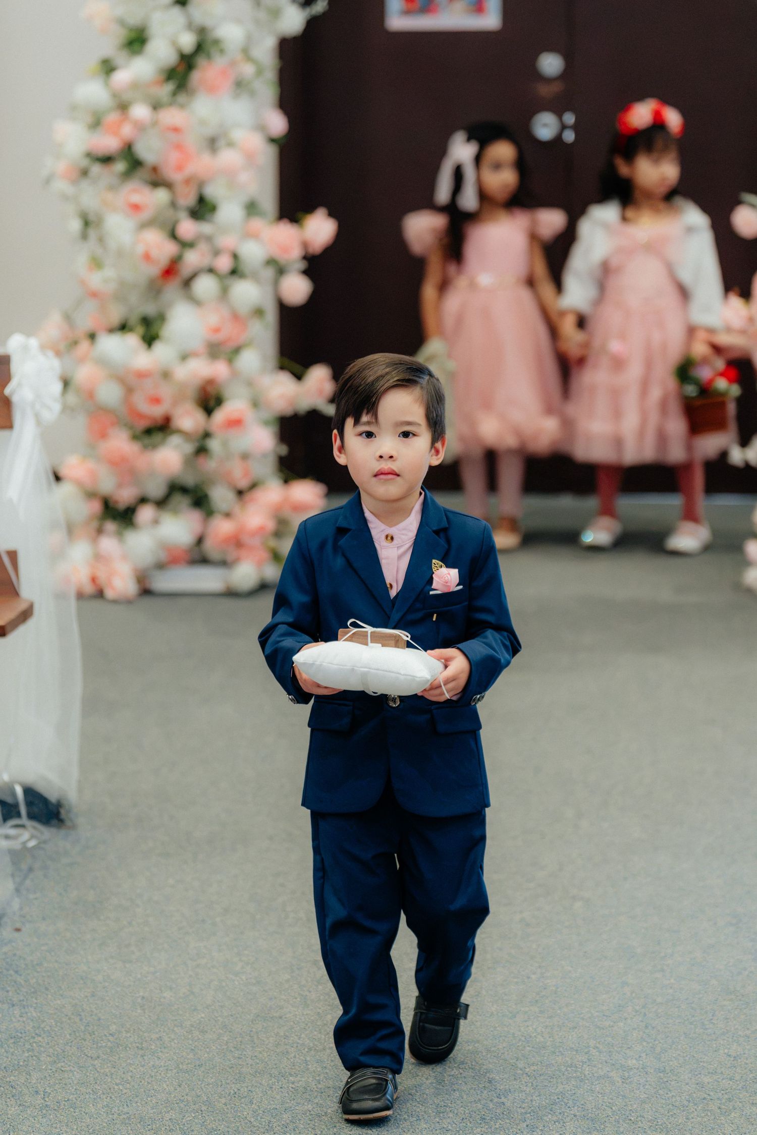 Young ring bearer in navy suit carrying white pillow walking down wedding ceremony aisle with flower girls.