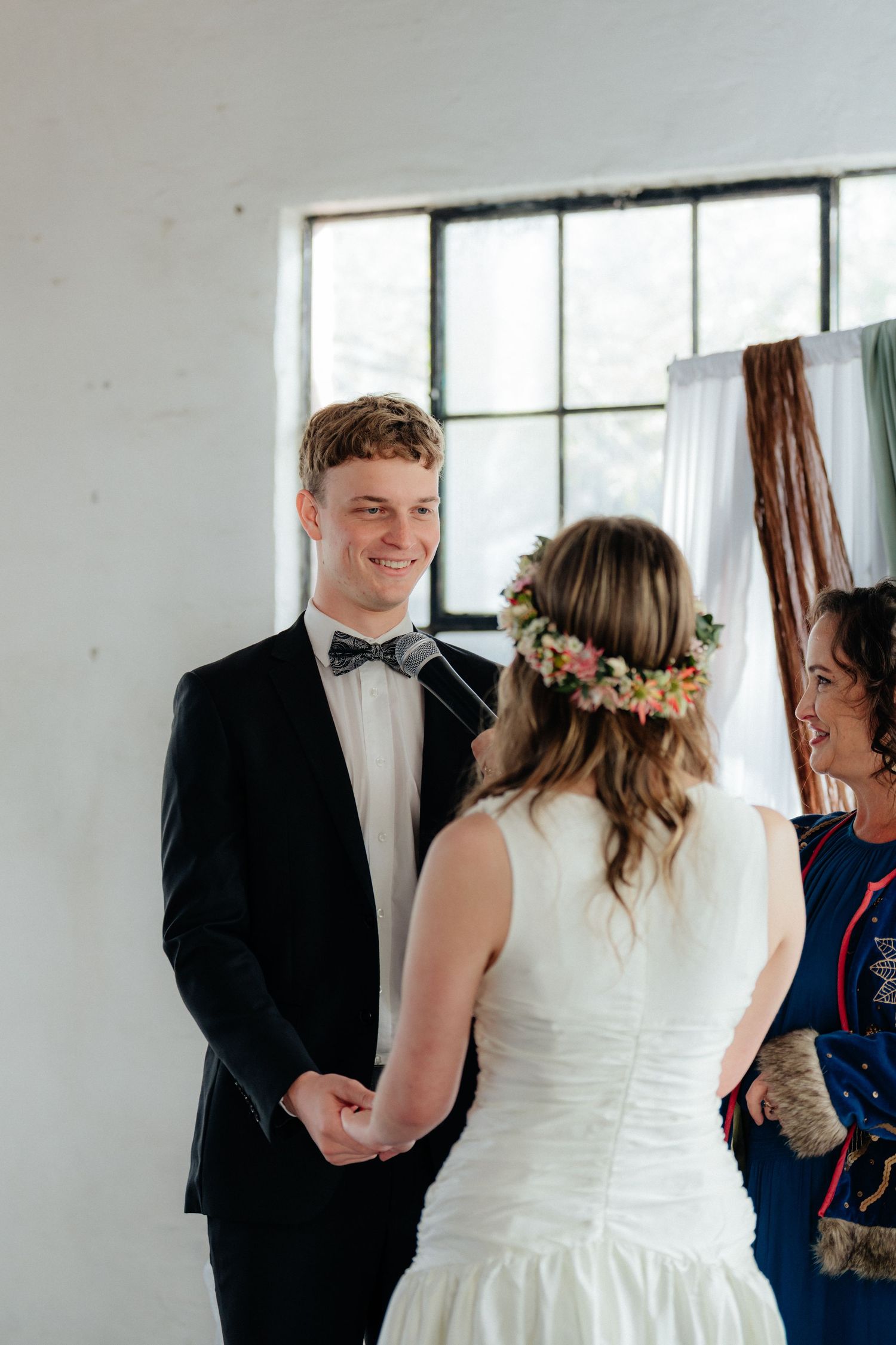 Smiling groom in black tuxedo holds brides hands during intimate indoor wedding ceremony.