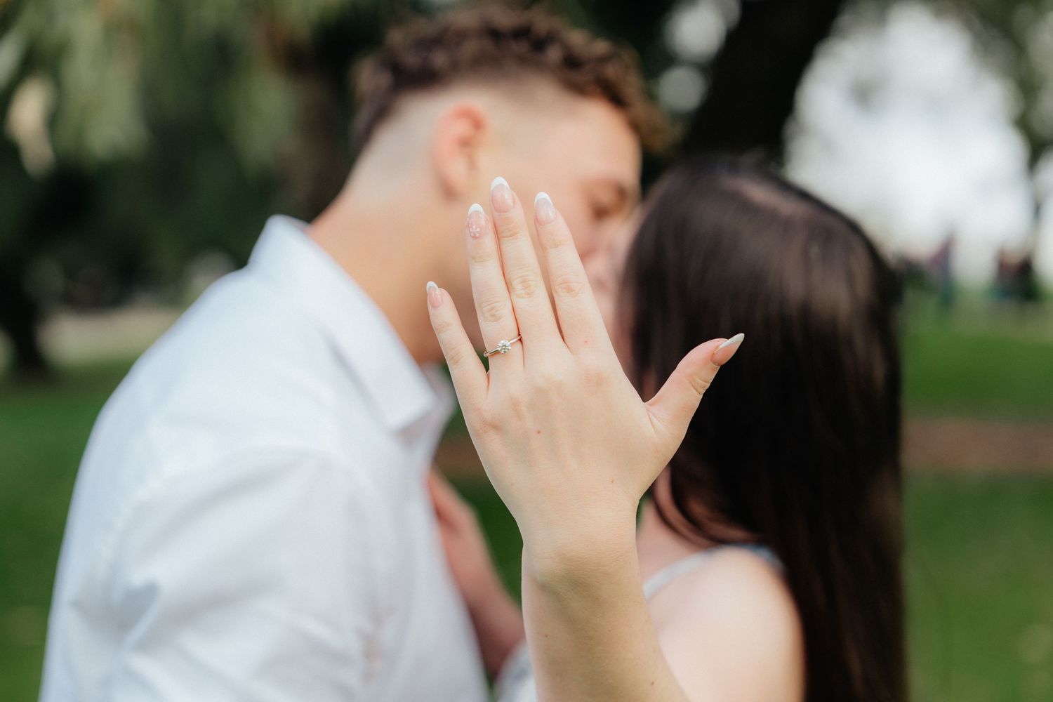 Engaged couple kissing while woman displays diamond engagement ring to camera outdoors.