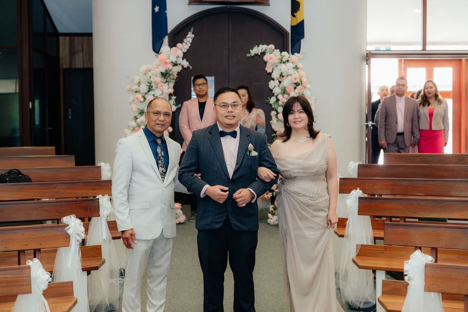 Groom escorted by parents down church aisle decorated with pink and white floral arrangements.